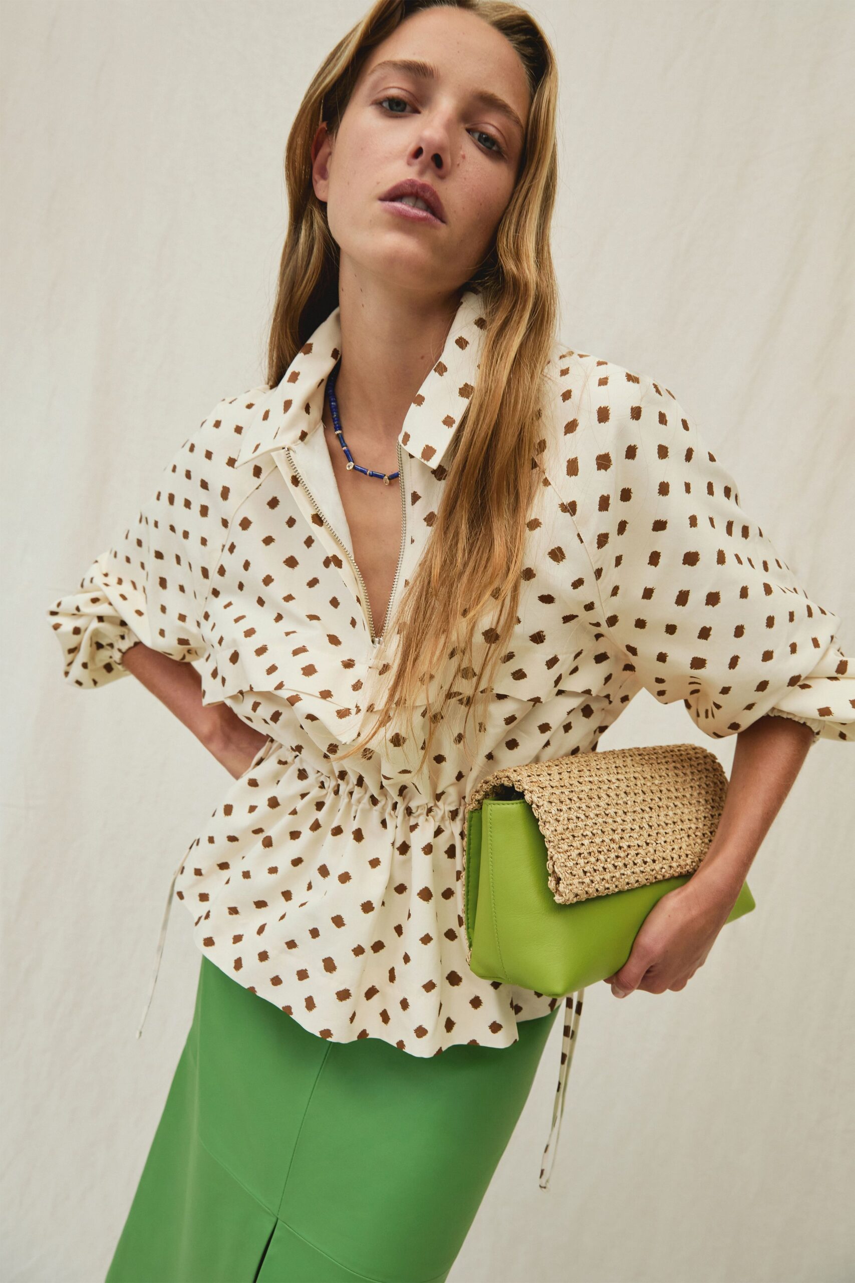 Cream zip-front blouse speckled in brown, drawstring waist cinched into a peplum. Short cobalt bead necklace with tiny pale spacers. Acid-green pencil skirt with a front slit and curved seam lines. Lime leather pouch tucked under a coarse raffia flap. Walking back from a beach lunch with sand still on the tablecloth.