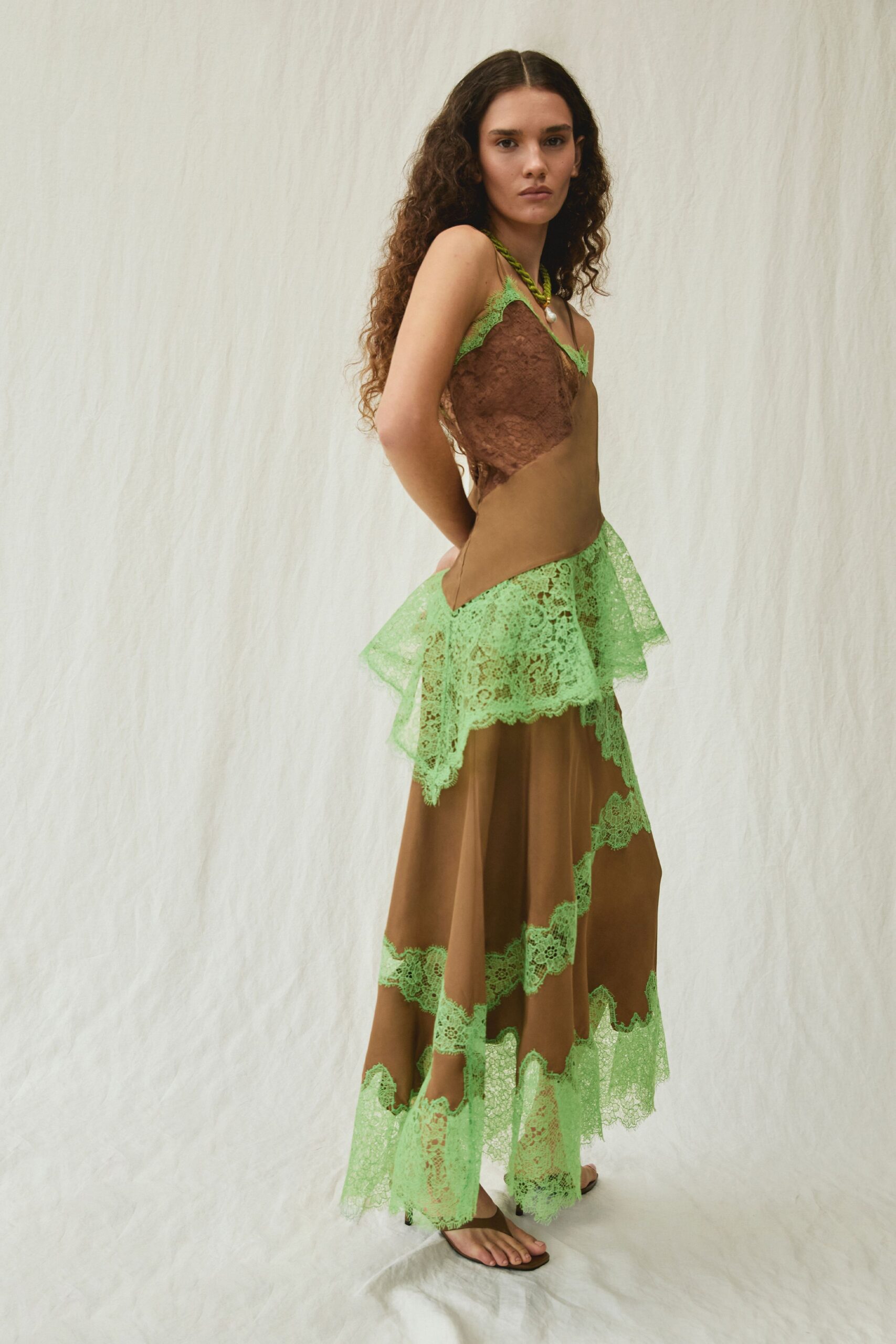 Bias-cut cocoa slip dress spliced with acid-lime lace flounces. Twisted green cord necklace with a single pearl drop. Flat brown thong sandals, barely there under the hem. Walking through a conservatory after rain, hem catching on damp stone.
