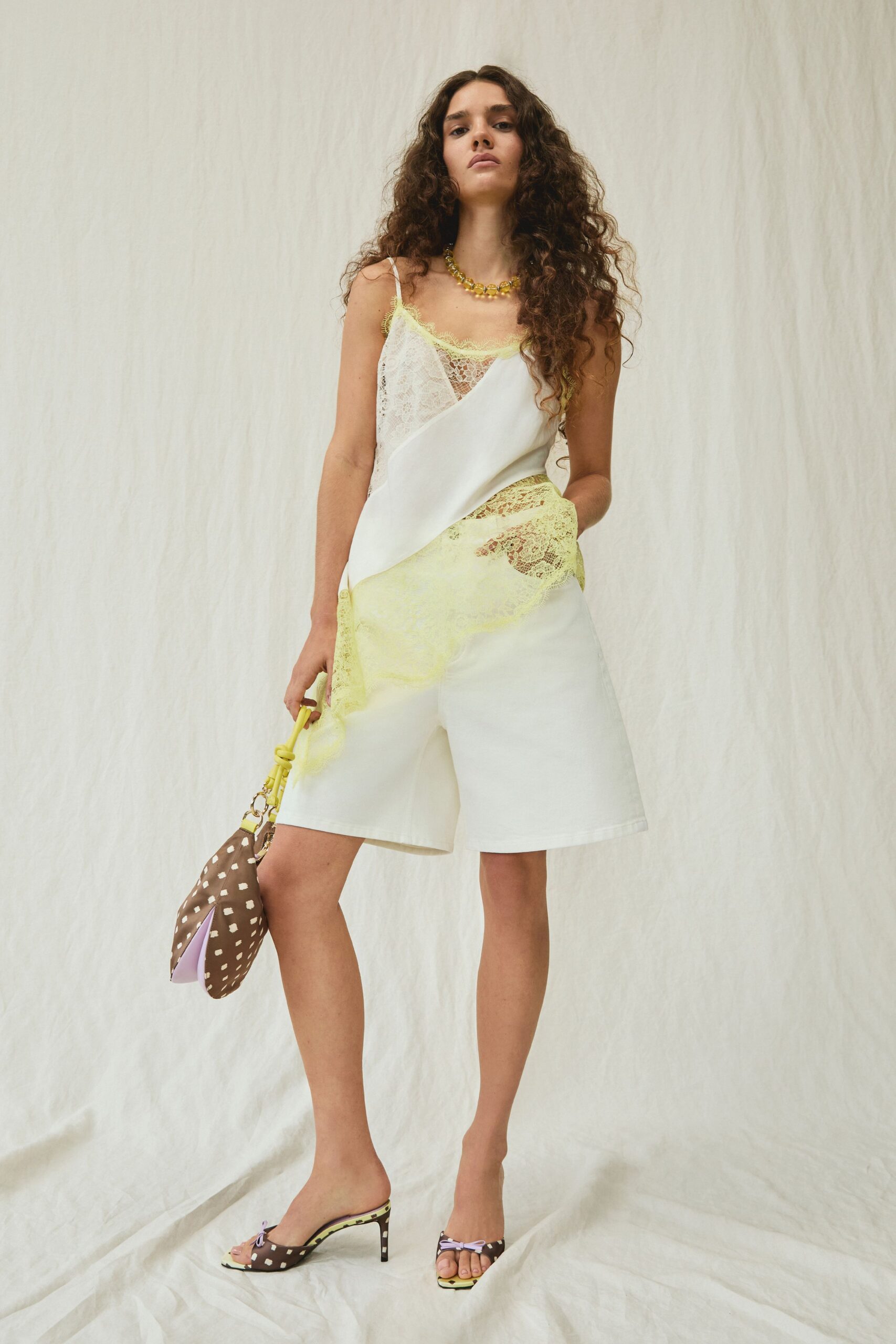 Ivory satin slip top sliced with panels of lemon eyelash lace. White tailored shorts, cut wide through the leg. Chunky amber-bead necklace at the collarbone. Brown pouch with scattered cream spots and a knotted strap. Brown spotted mules with lilac bow toes. Walking back from a garden lunch with pears in the bag.