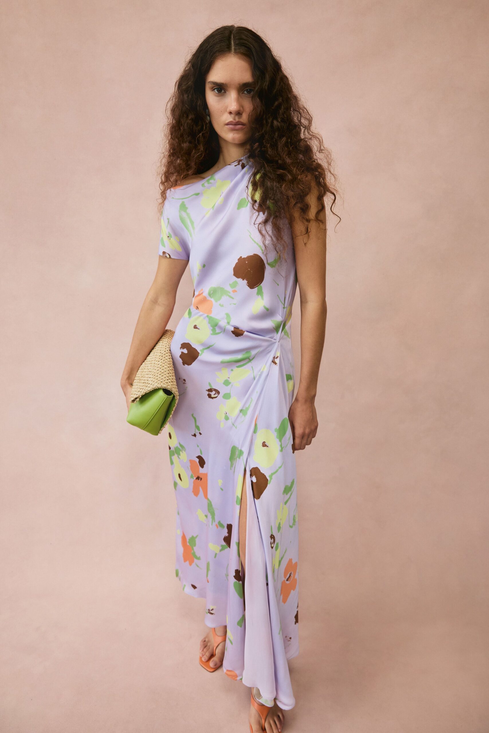 Lavender jersey column dress, one-shoulder drape twisted tight at the hip slit. Woven straw foldover clutch with a lime leather base peeking out. Apricot thong sandals, square-toed and nearly flat. Walking back from a seaside lunch with salt drying on your shoulders.