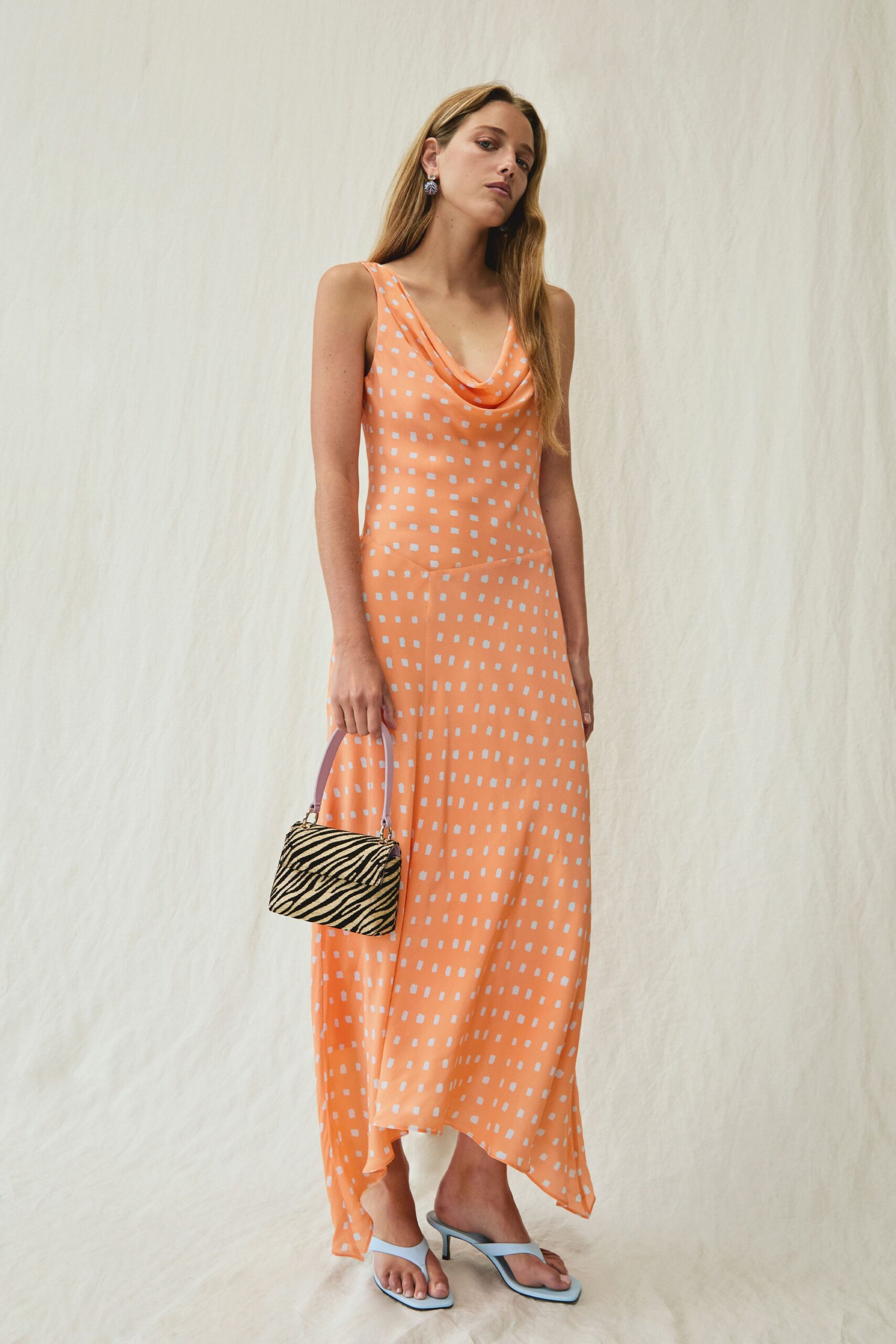 Peach slip dress in fluid satin, white dash print and a cowl neck. Small clustered drop earrings. Boxy zebra bag with a lilac handle. Pale blue thong heels with square toes. Walking back from a beach wedding after the music has ended.