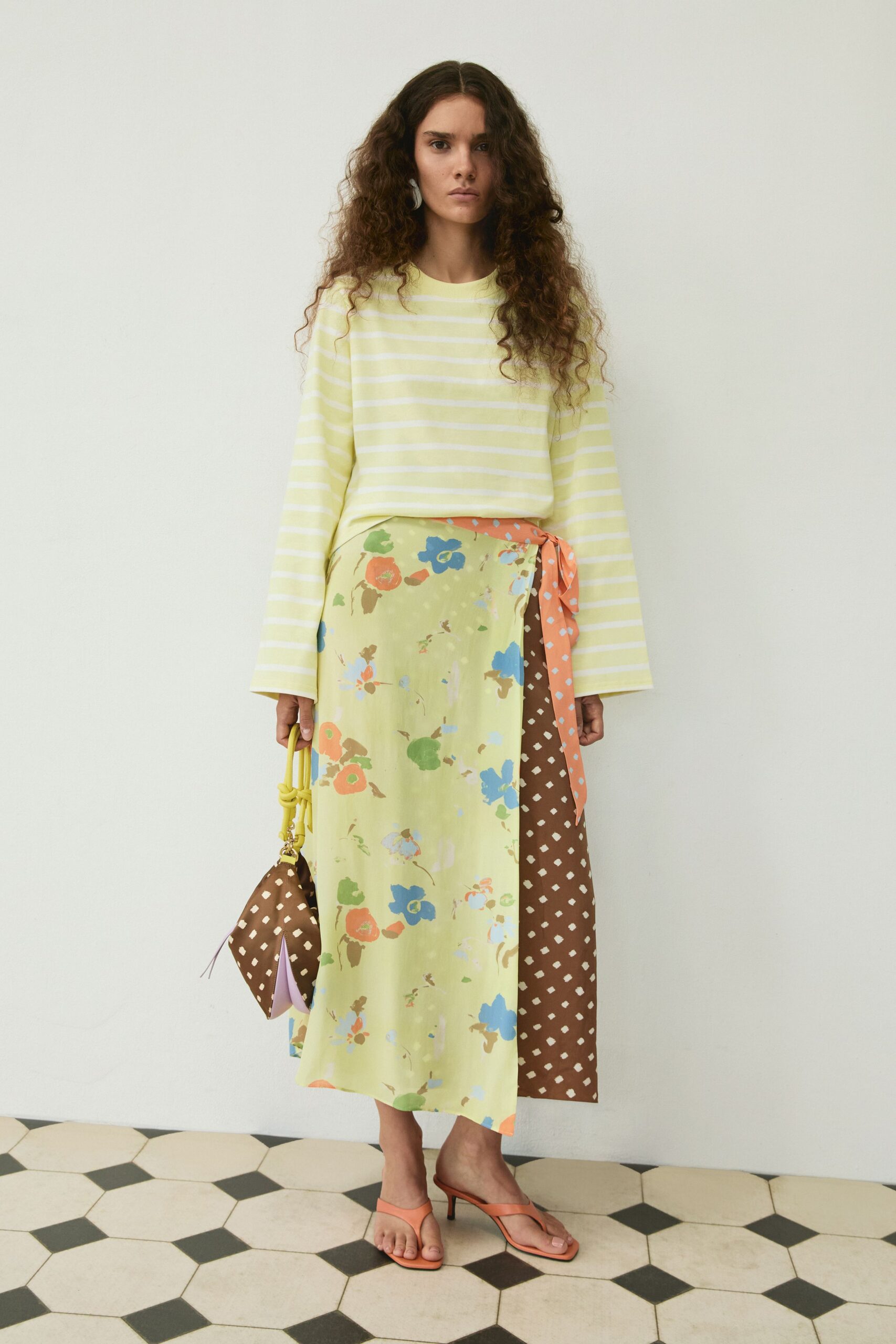 White curved drop earrings. Pale lemon striped knit top with long sleeves flaring at the cuff. Bias-cut midi wrap skirt splicing watery florals with brown diamond print. Brown pouch with a knotted yellow handle. Peach thong mules on a low kitten heel. Walking through a tiled courtyard after a long seaside lunch.