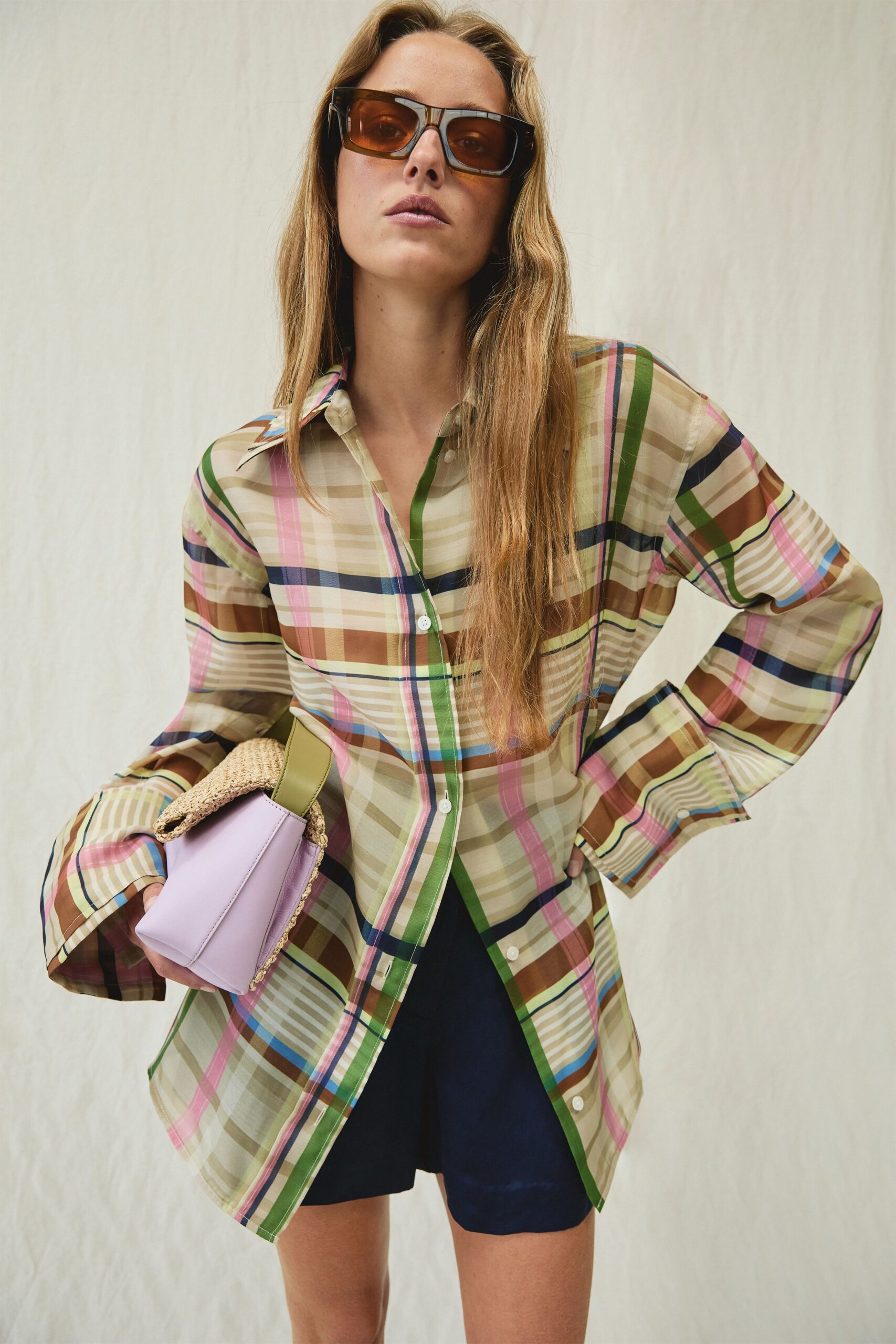 Oversized square frames with amber lenses in glossy tortoiseshell. Sheer beige plaid shirt with green and pink stripes, one button skewed open. Dark navy satin shorts cut loose and high on the thigh. Lilac leather box bag capped with a folded raffia flap. Walking back from a resort breakfast with the room key still missing.
