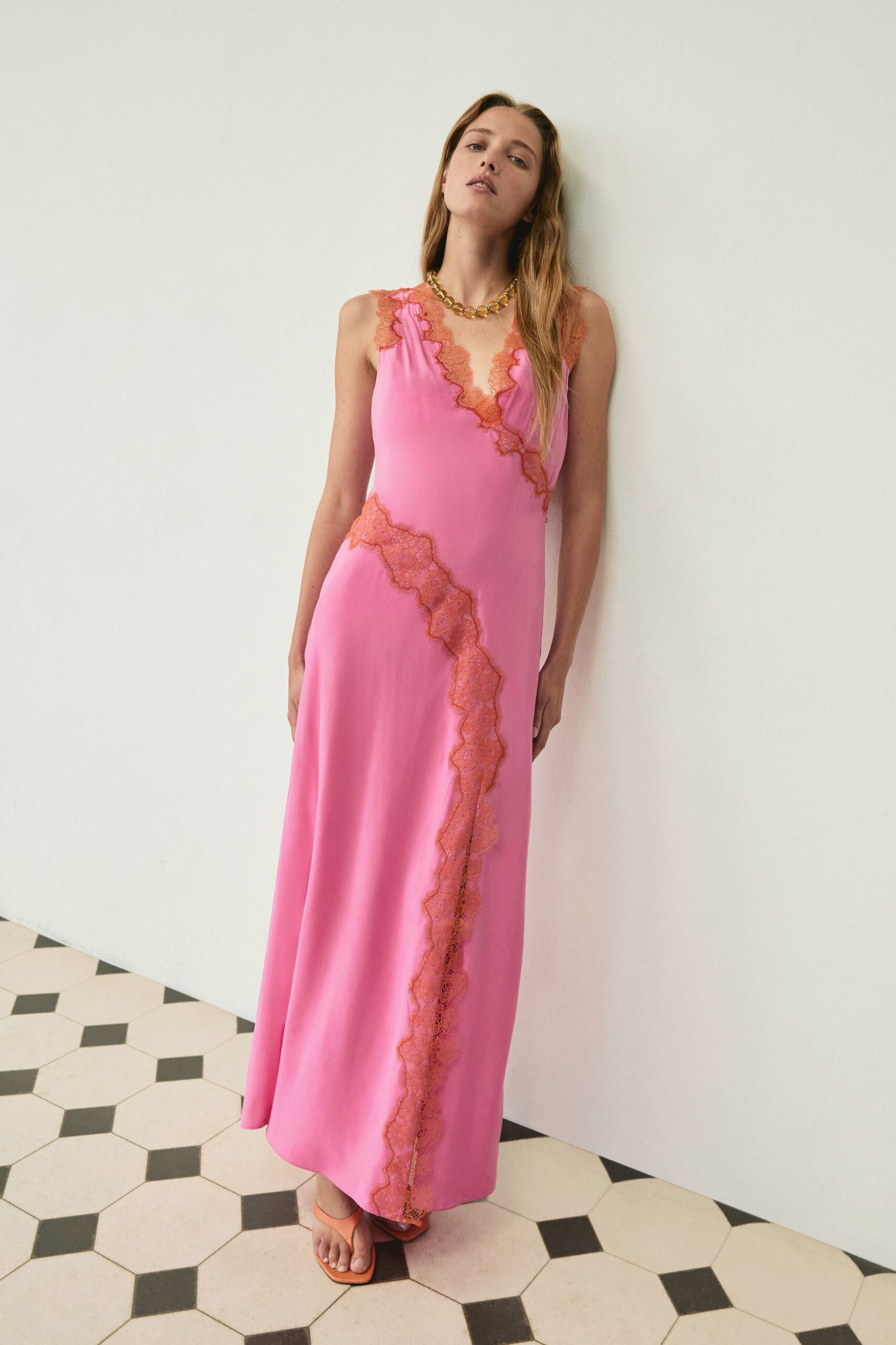 Bubblegum pink slip dress with rust lace spliced diagonally through the skirt. Chunky amber-toned resin links at the collarbone. Peach square-toe sandals, one foot edging out at the hem. Hotel corridor afternoon, tiles still cool after the mop bucket passed.