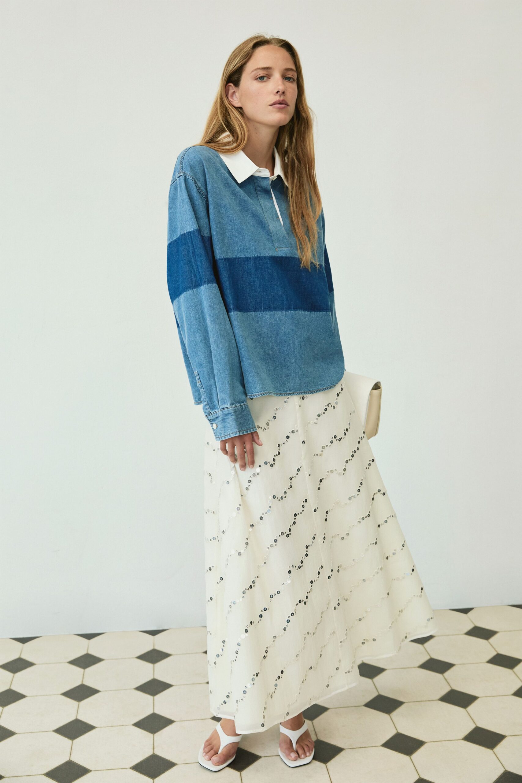 Chambray polo in three washed indigo bands with a stark white collar. Ivory midi skirt scattered with diagonal rows of tiny silver sequins. Cream foldover clutch tucked flat under the arm. White thong sandals with squared soles. Hotel breakfast after a swim, tiles still cold underfoot.