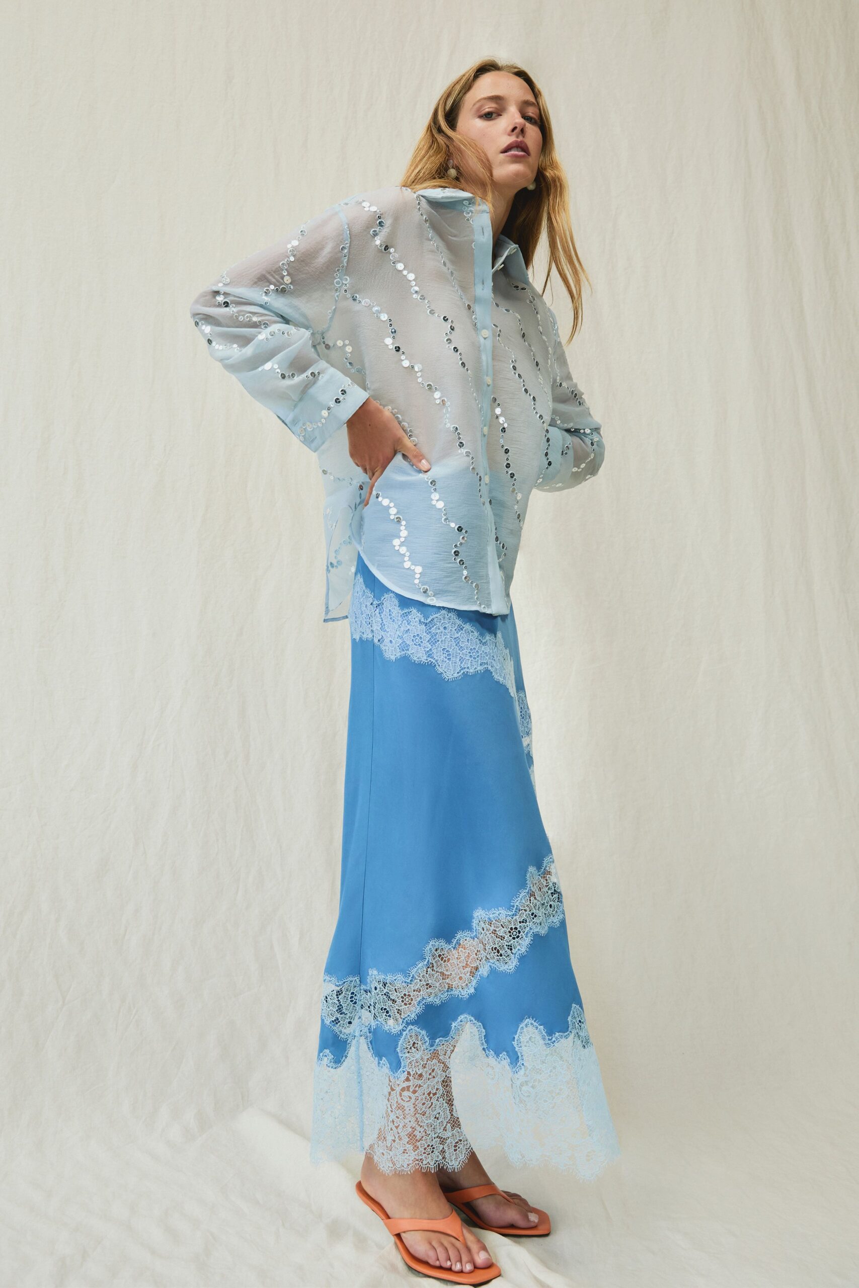 Sheer ice-blue shirt traced with wavy rows of silver sequins. Cornflower satin slip skirt banded with white eyelash lace at hip and hem. Apricot thong sandals with squared soles. Walking out to a hotel terrace just after the champagne tower collapsed.