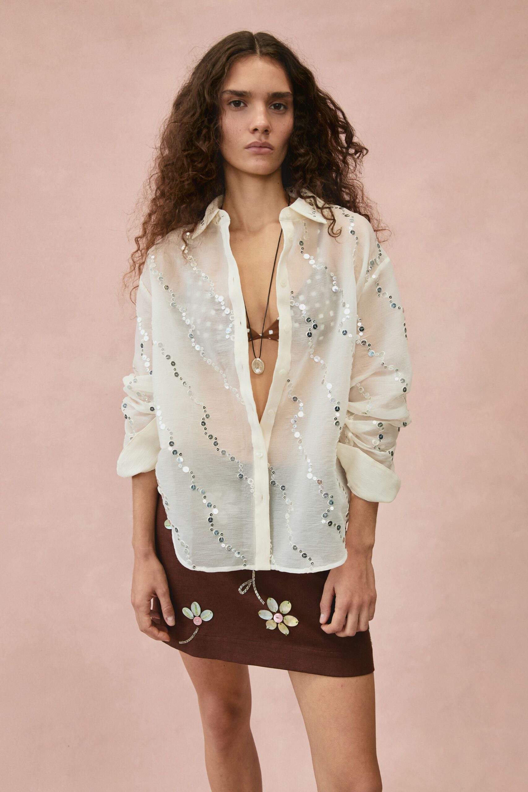Sheer ivory shirt traced with winding rows of silver sequins. Chocolate triangle bralette, barely held by thin cord straps. Small oval pendant on a long black thong. Brown mini skirt appliqued with mother-of-pearl daisies near the hem. Walking back from the beach bar before the tablecloths come off the terrace.