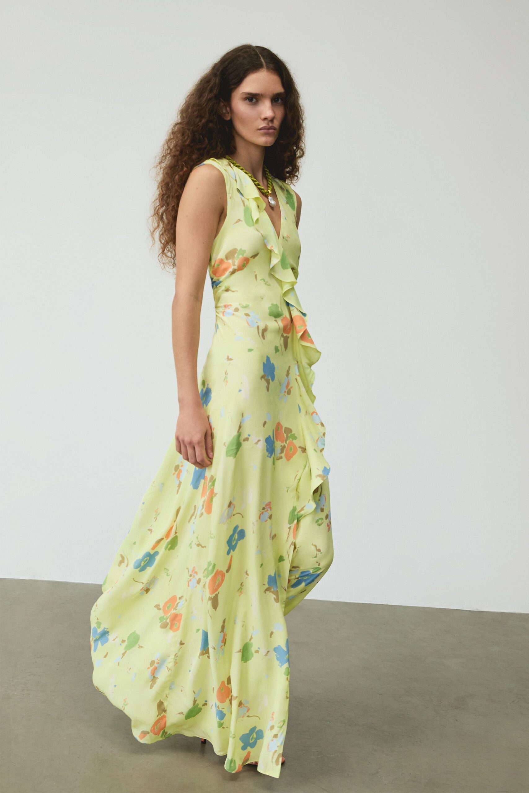 Twisted green cord necklace with a single baroque pearl drop. Butter-yellow sleeveless dress in fluid satin, ruffle running off-center through the floral print. Garden wedding spillover, someone has opened another bottle on the lawn.