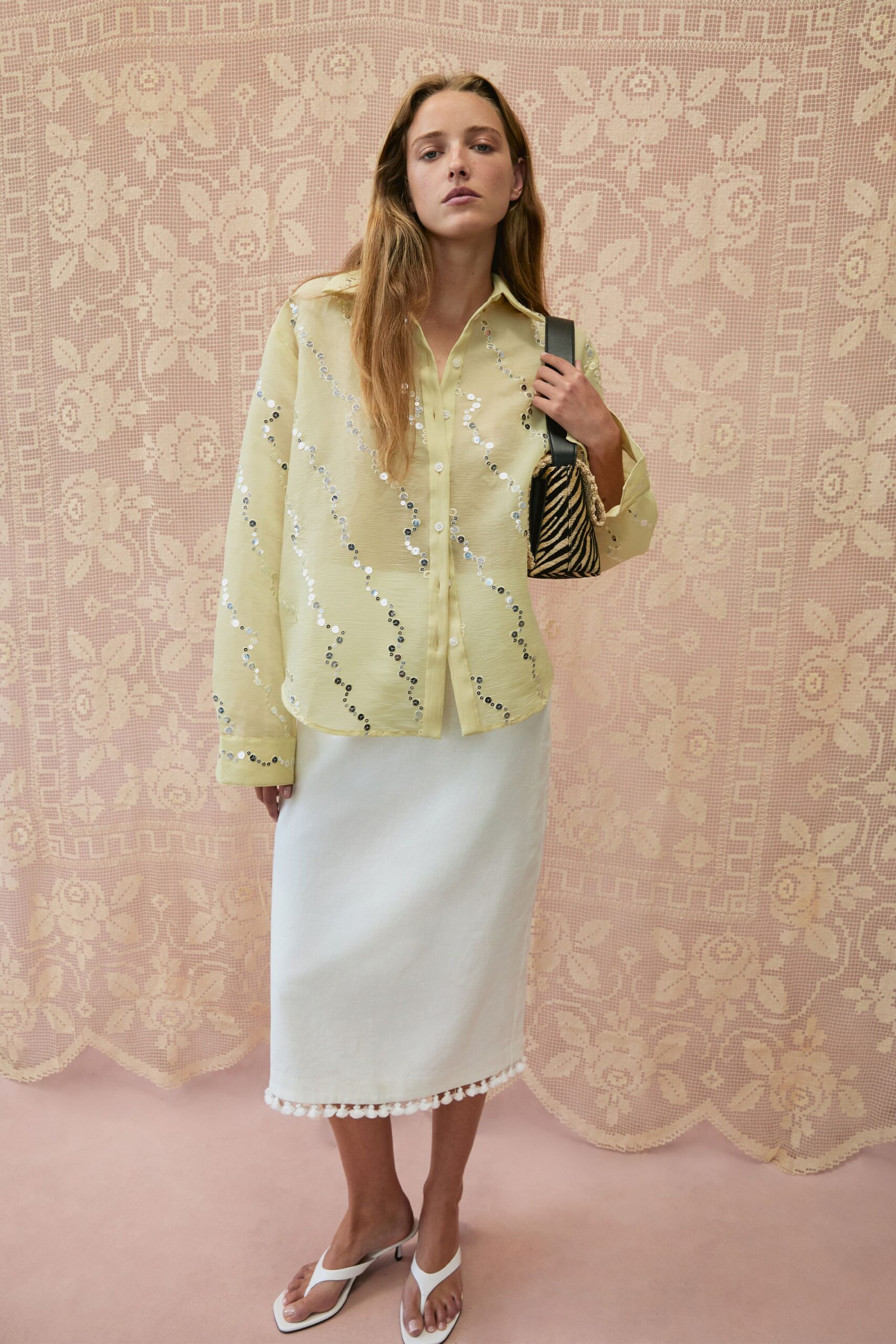 Butter-yellow crepe shirt traced with silver sequins in loose vertical waves. Small zebra pouch on a wide black strap, rope tassel at the side. Ivory column skirt with a pompom fringe tapping the hem. White thong heels with square toes. Sunday hotel terrace after the pool, still carrying someone else's room key.