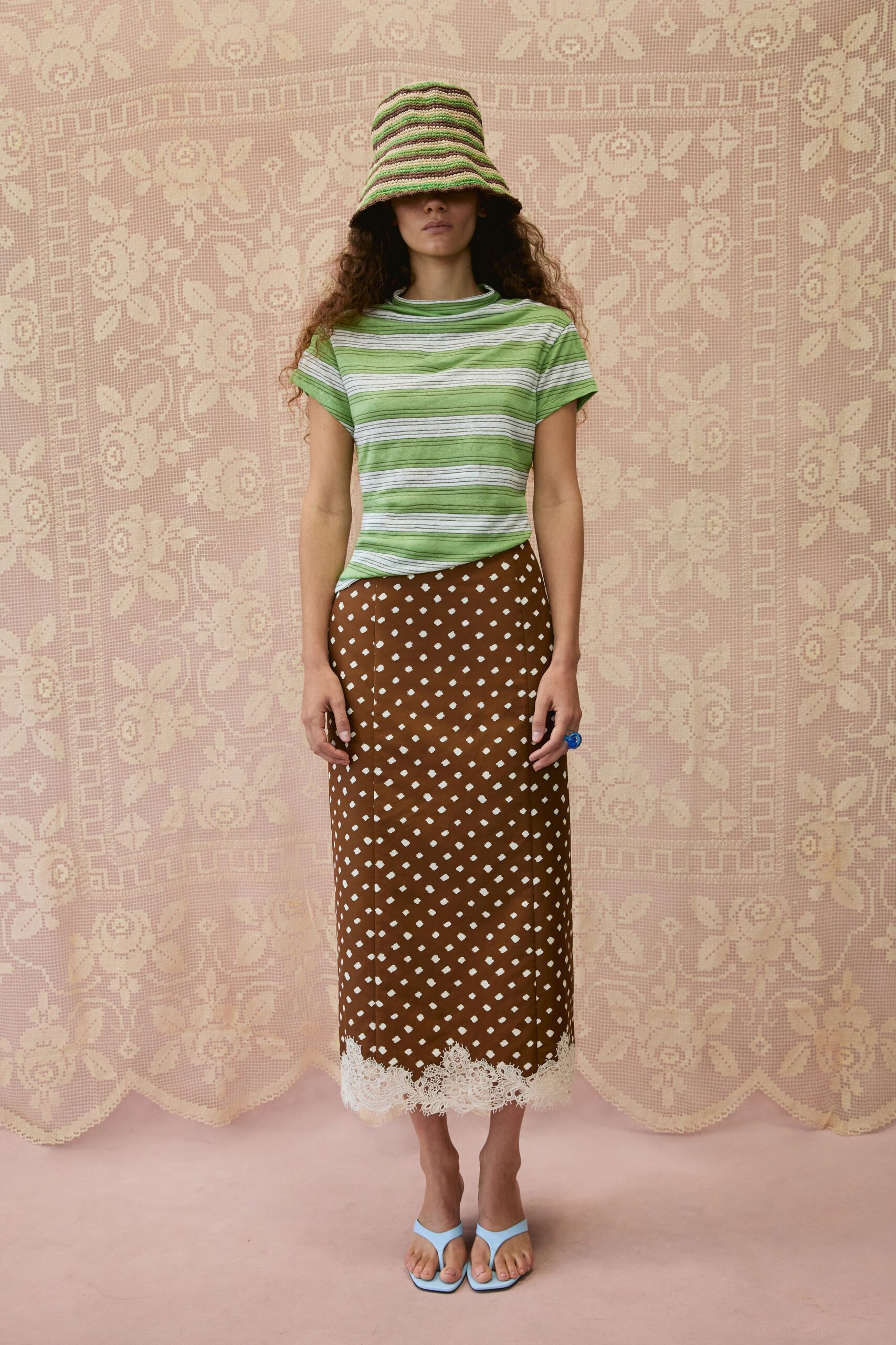 Crocheted bucket hat in brown, cream and mint stripes, brim hiding the eyes. Green and white striped knit tee with rolled cap sleeves. Brown column skirt scattered with white dots, eyelash lace at the hem. Chunky cobalt resin ring. Powder-blue thong sandals. Sunday cake table under a lace cloth, shoes kicked off in the grass.