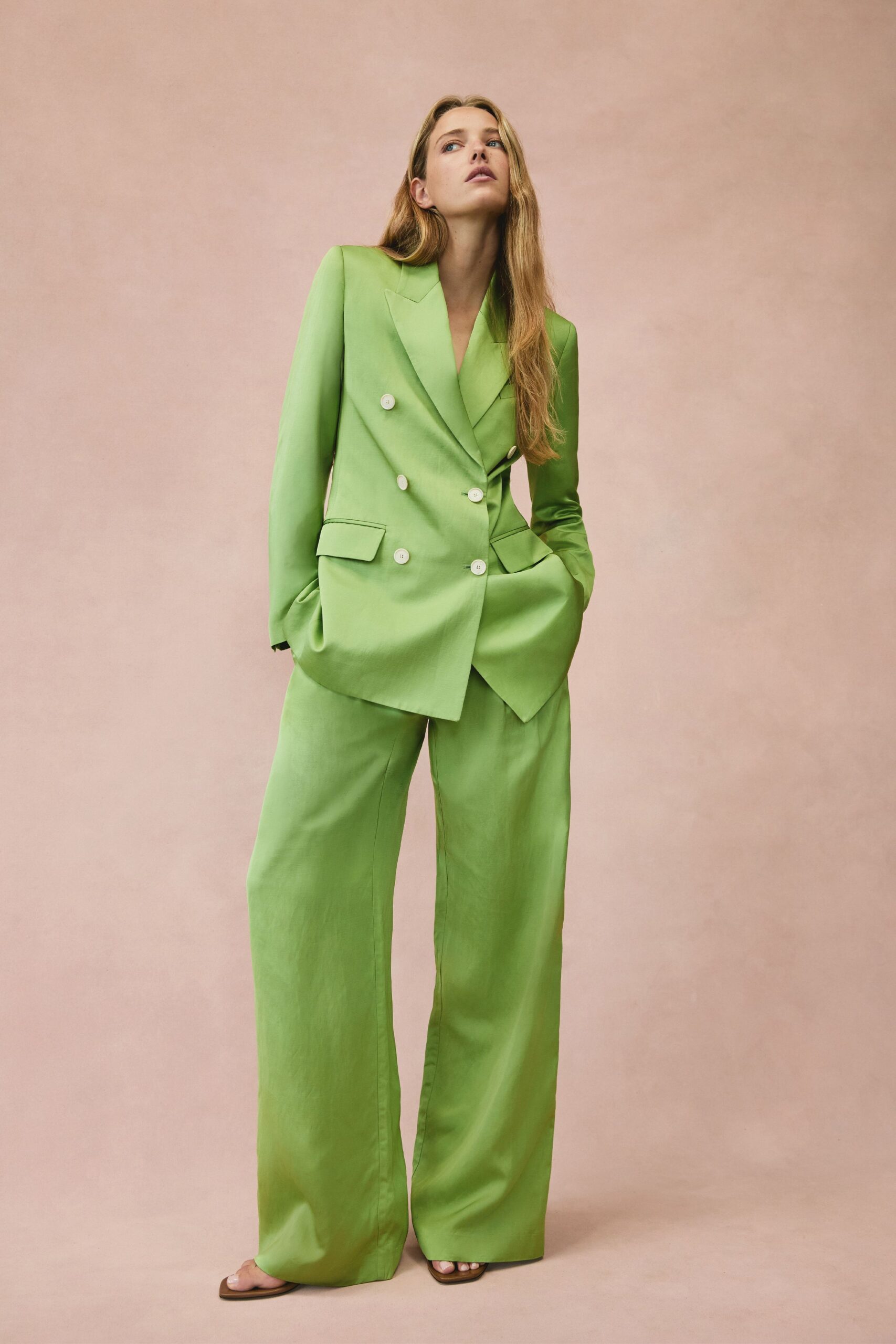 Lime double-breasted blazer with sharp peak lapels and four ivory buttons. Matching lime trousers in fluid suiting, hems pooling over the sandals. Flat tan sandals with squared toes peeking out. For opening windows in a rented apartment on the first warm day.