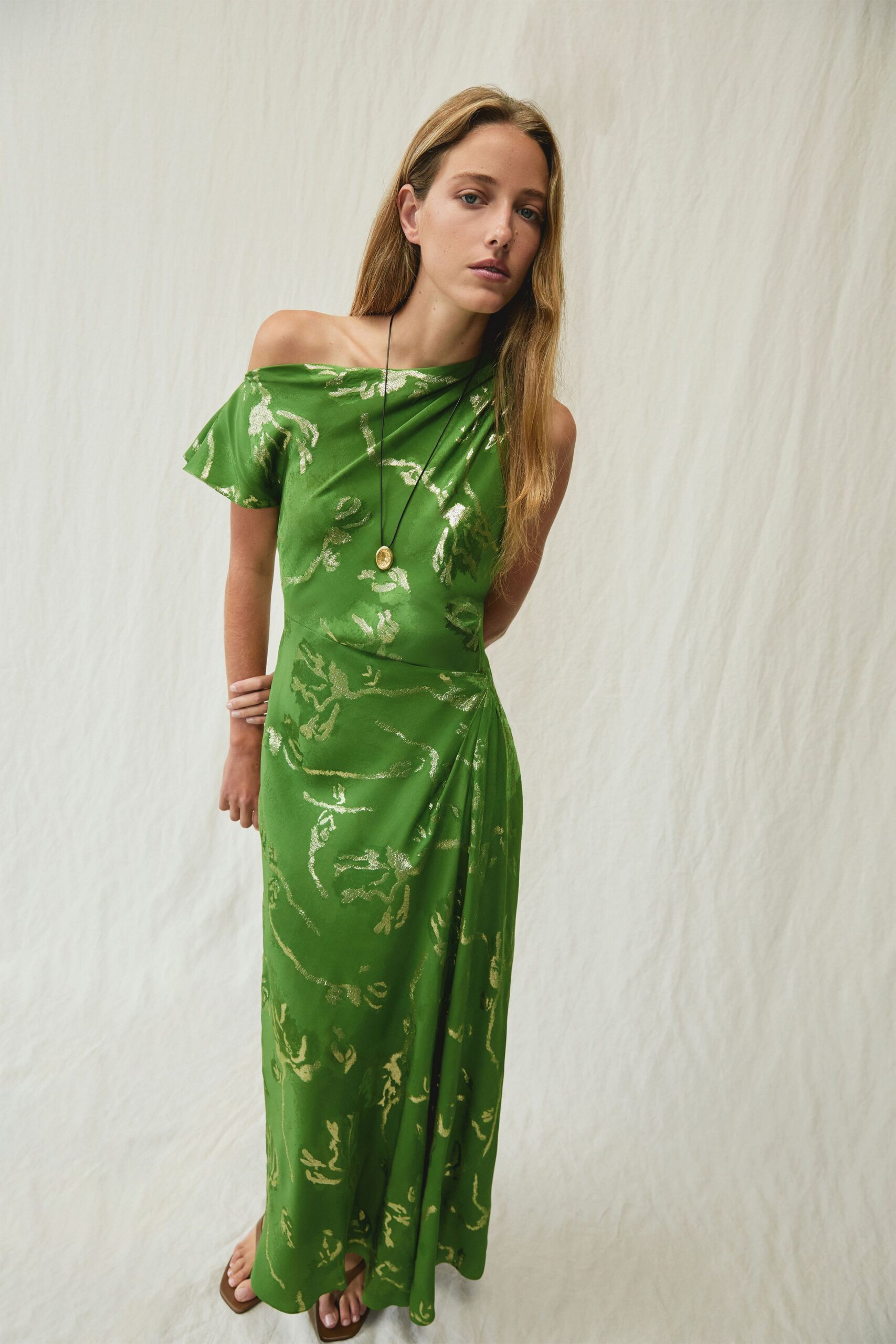 Grass-green draped dress with a one-shoulder neckline and pale metallic vine jacquard. Small oval medallion hanging low on a black cord. Flat brown sandals with squared soles peeking past the hem. Terrace wedding heat, someone has moved the chairs into the shade.