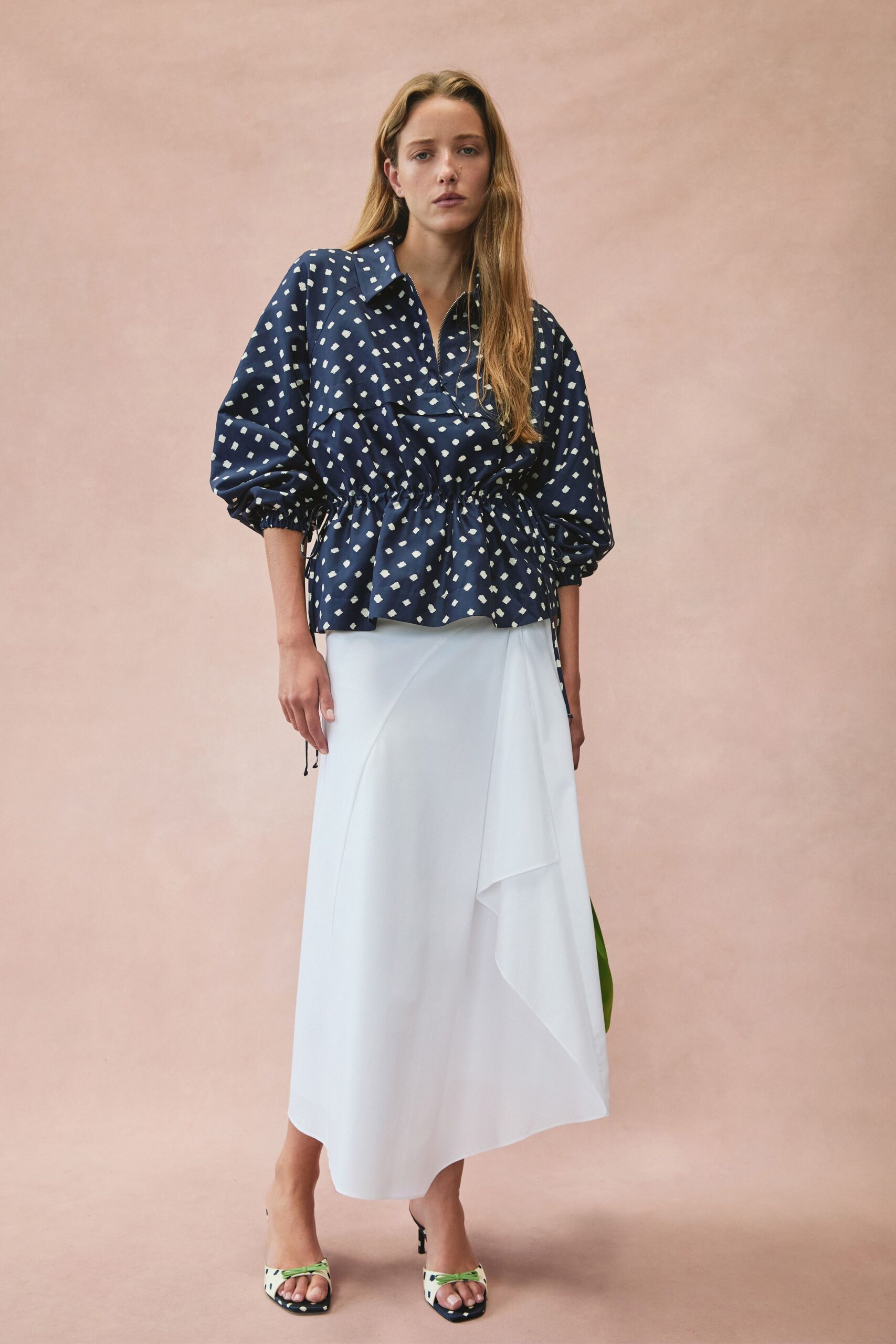 Navy blouse scattered with chalk square dots, drawstring waist puckering the peplum. White wrap skirt in fluid crepe, one panel falling in a sharp cascade. Mismatched print mules with lime knotted straps across the toes. For lunch on a hotel terrace once the sea wind starts lifting napkins.