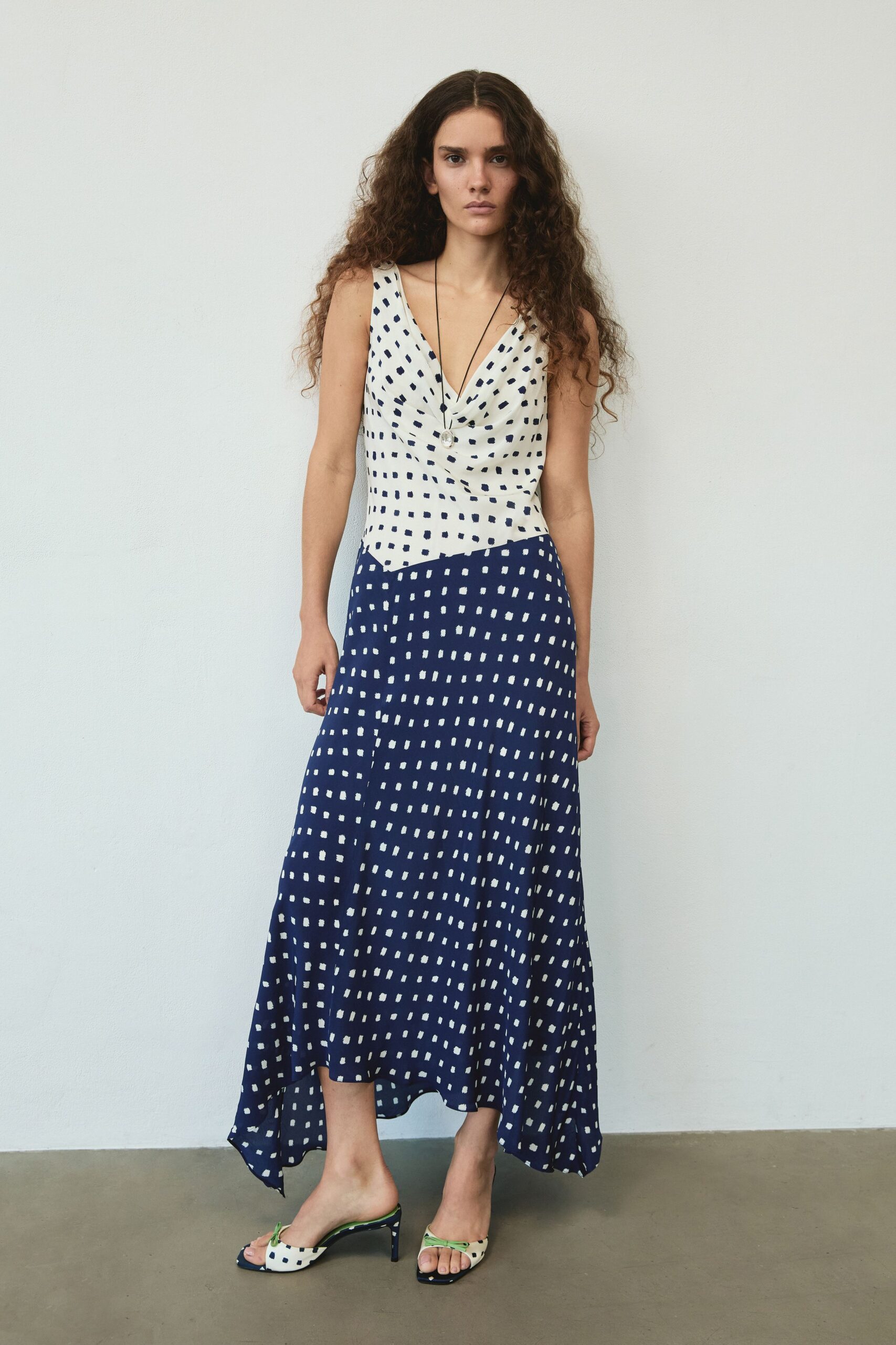Long black cord with a cloudy oval pendant at the sternum. Ivory-to-navy bias dress, draped cowl neckline and scattered brushmark dots. Open-toe kitten-heel mules with mismatched dotted panels and lime straps. Walking across a tiled courtyard after lunch, heat still coming off the walls.