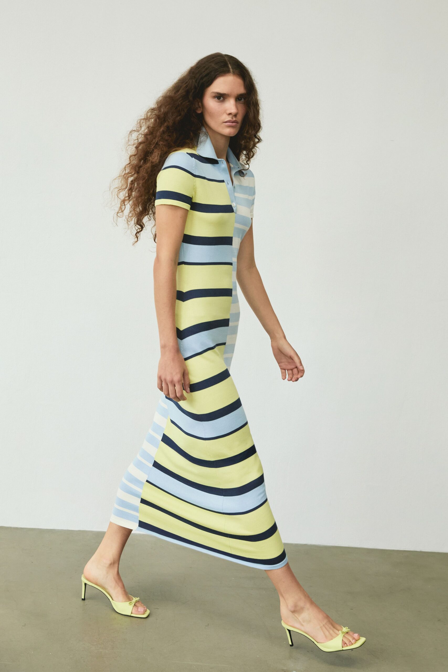 Body-skimming knit polo dress, lemon and powder-blue stripes switching direction at the placket. Pale chartreuse mule sandals with a tiny bow pinching the toe strap. Walking the marina promenade before the wind gets up.