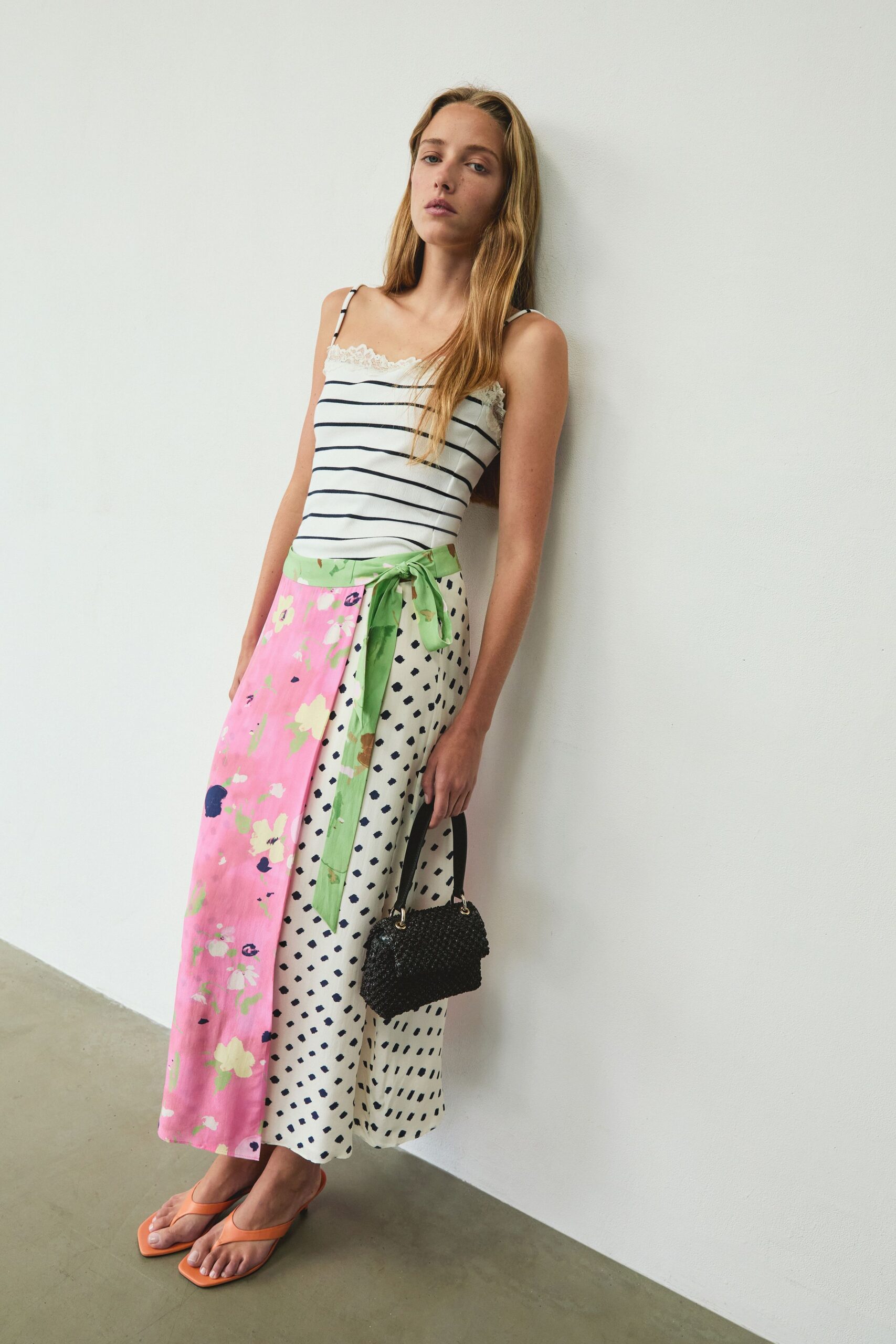 White striped camisole with scalloped lace peeking above the straight neckline. Midi wrap skirt split between pink florals and navy dash print, green sash tied loose. Black beaded mini bag with silver ring hardware. Apricot thong sandals on a low flare heel. Sunday courtyard brunch, somebody brought the good peaches.