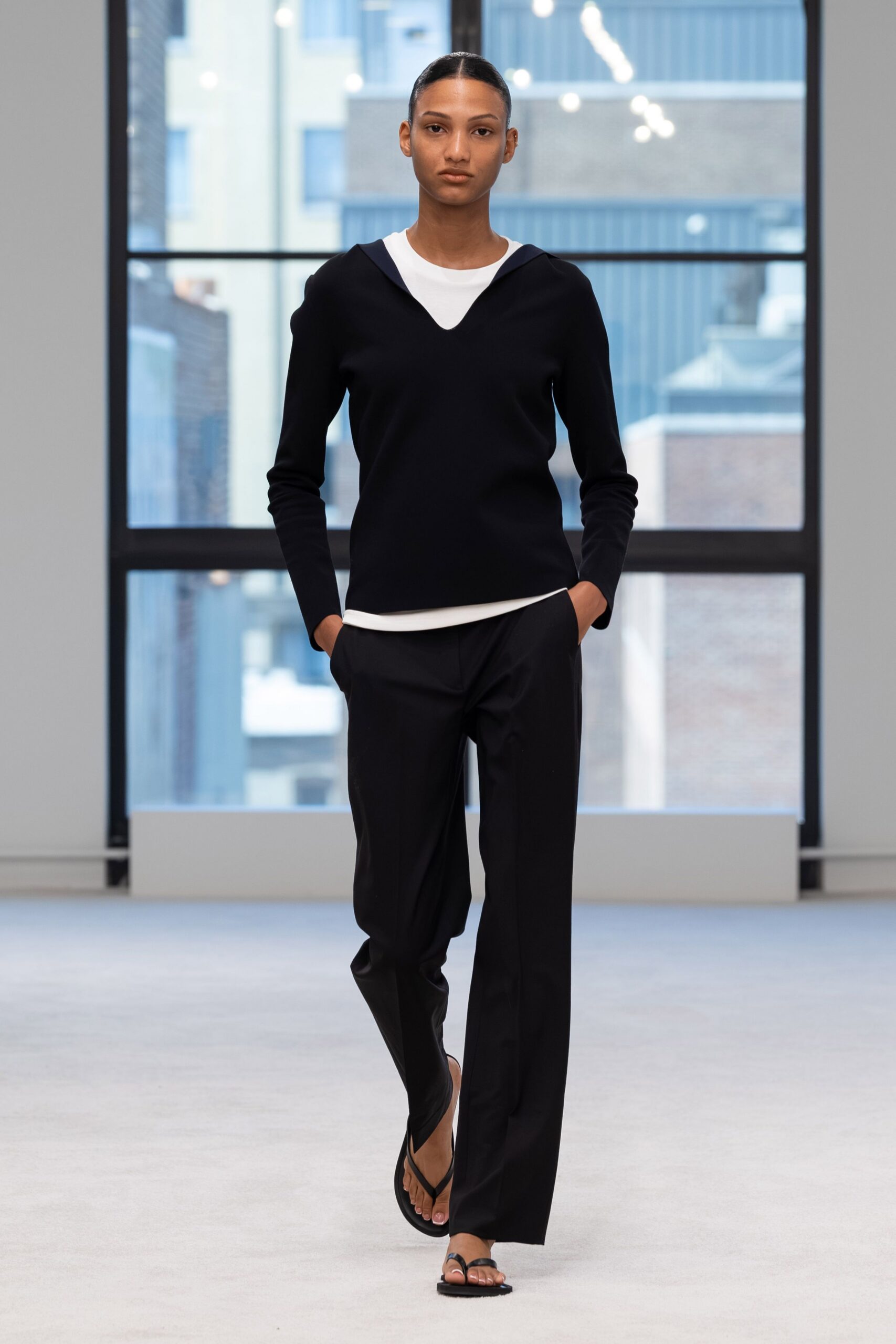 Black fine-knit top with a sailor collar and dipped V opening. White jersey crewneck peeking out in a clean crescent. Black straight trousers in fluid suiting cloth, pressed through the leg. Flat thong sandals with a slim toe loop. Early ferry crossing, windows steamed and the coffee already gone.