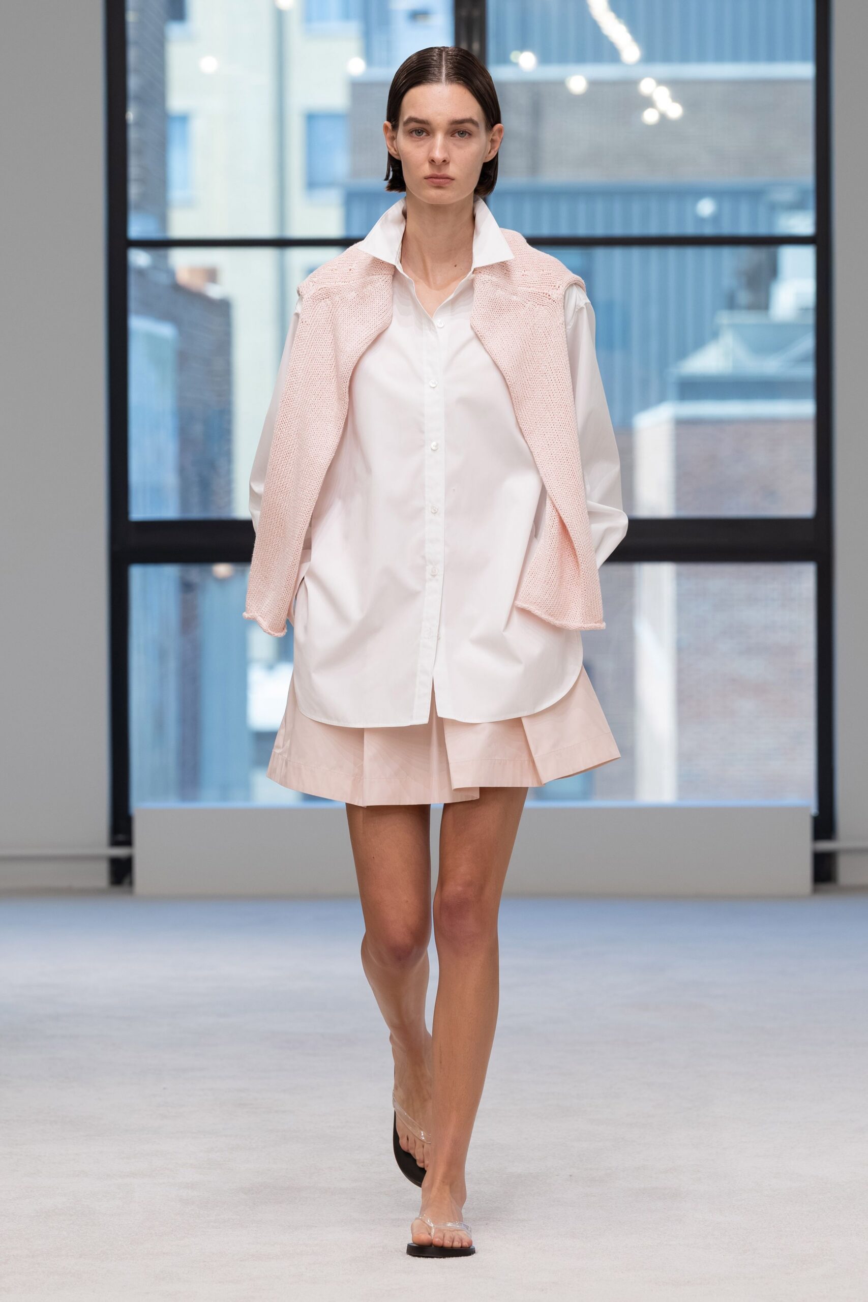 Pale blush open-knit sweater draped over the shoulders, rolled edges curling. Long white poplin shirt with a high spread collar and low hem. Blush mini skirt with a crisp flared panel peeking below. Clear vinyl thong sandals with square black soles. Office tower atrium, the first warm morning after keeping a coat on too long.
