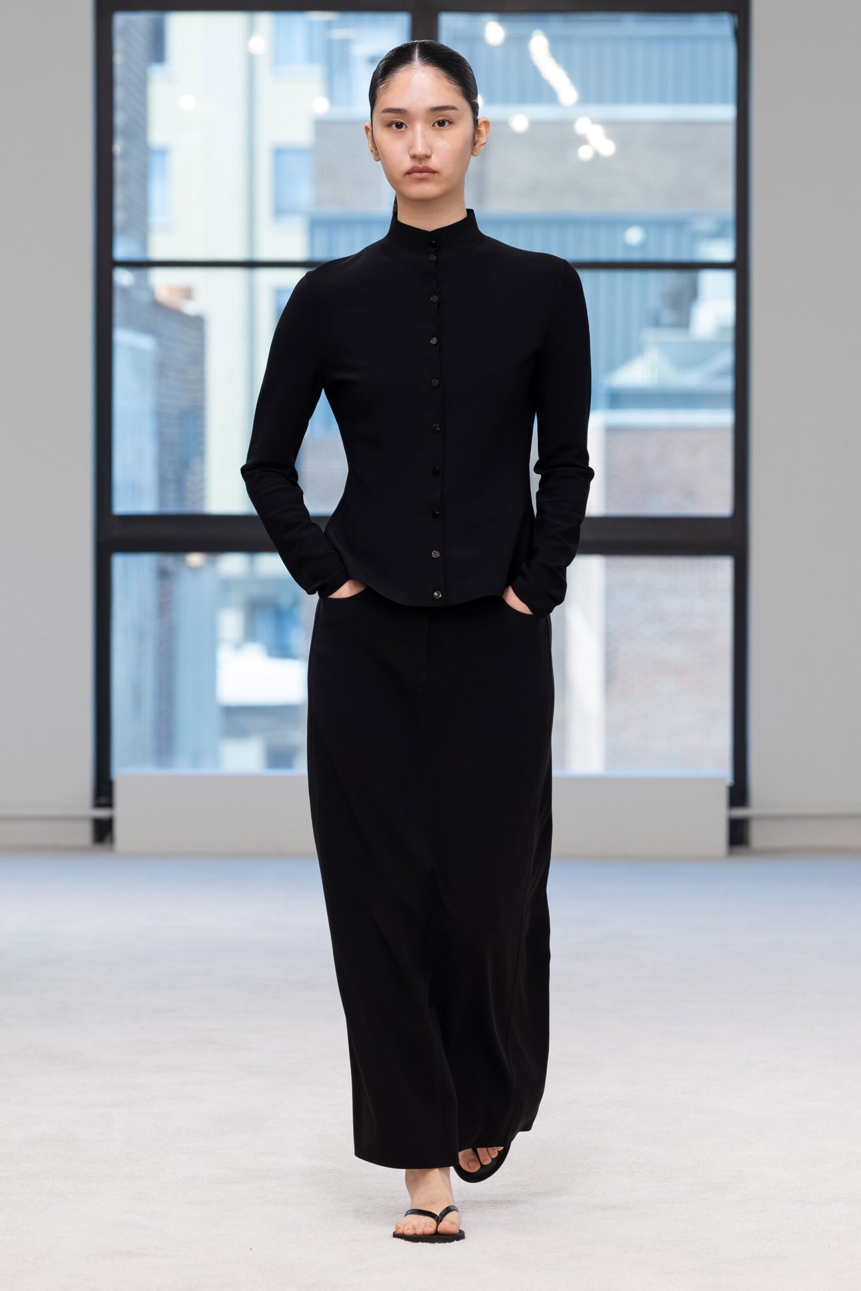 Black fine-gauge knit cardigan, stand collar and tiny tonal buttons. Floor-skimming black knit column, pockets opening at the hip. Flat black thong sandals. First morning in a borrowed apartment, city roofs still blue with rain.