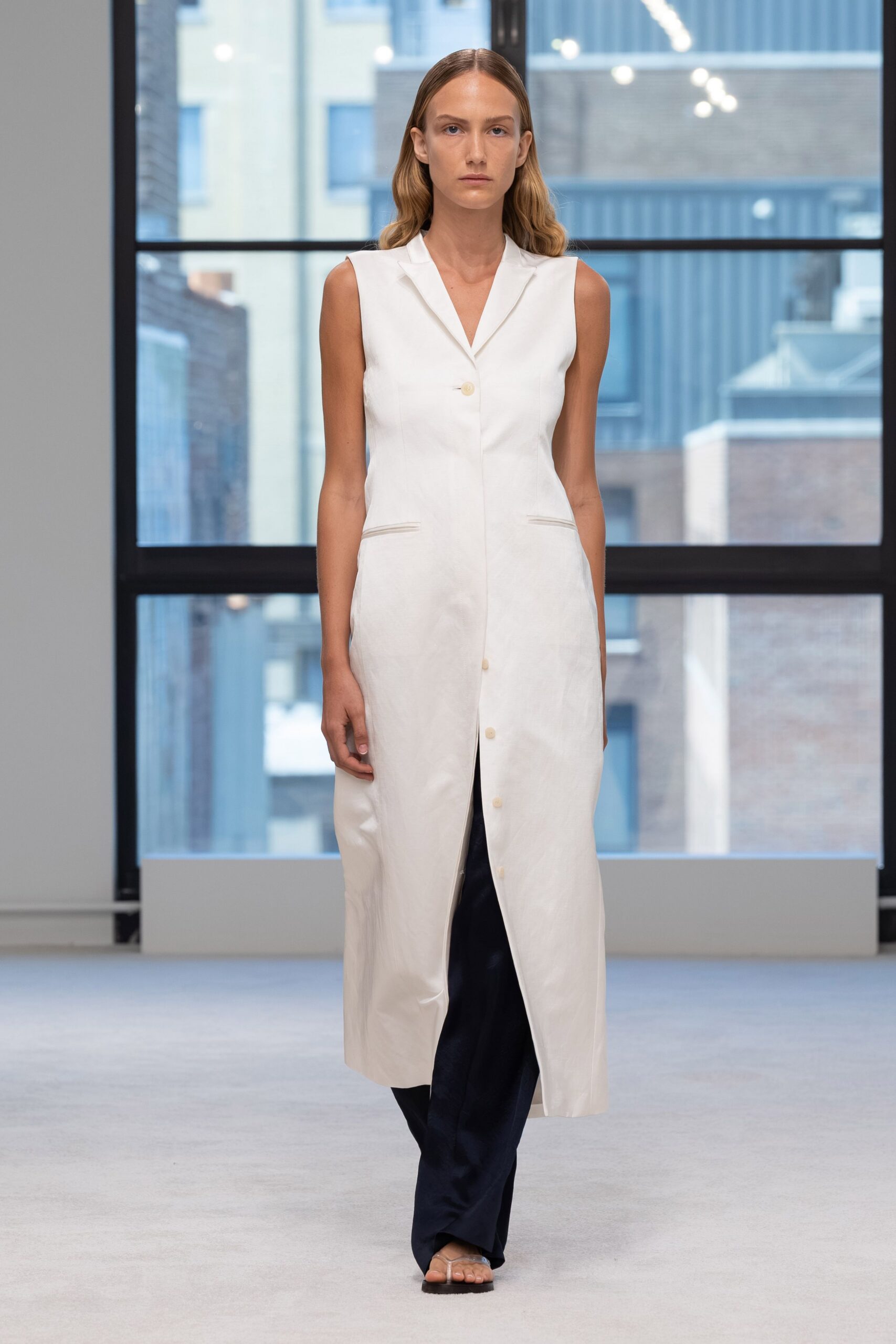 Sleeveless ivory coat-dress in crisp satin, notched lapels and low welt pockets. Ink wide-leg trousers breaking softly through the front slit. Clear thong sandals on a dark flat sole. Early meeting in a glass tower before the cleaners have finished.