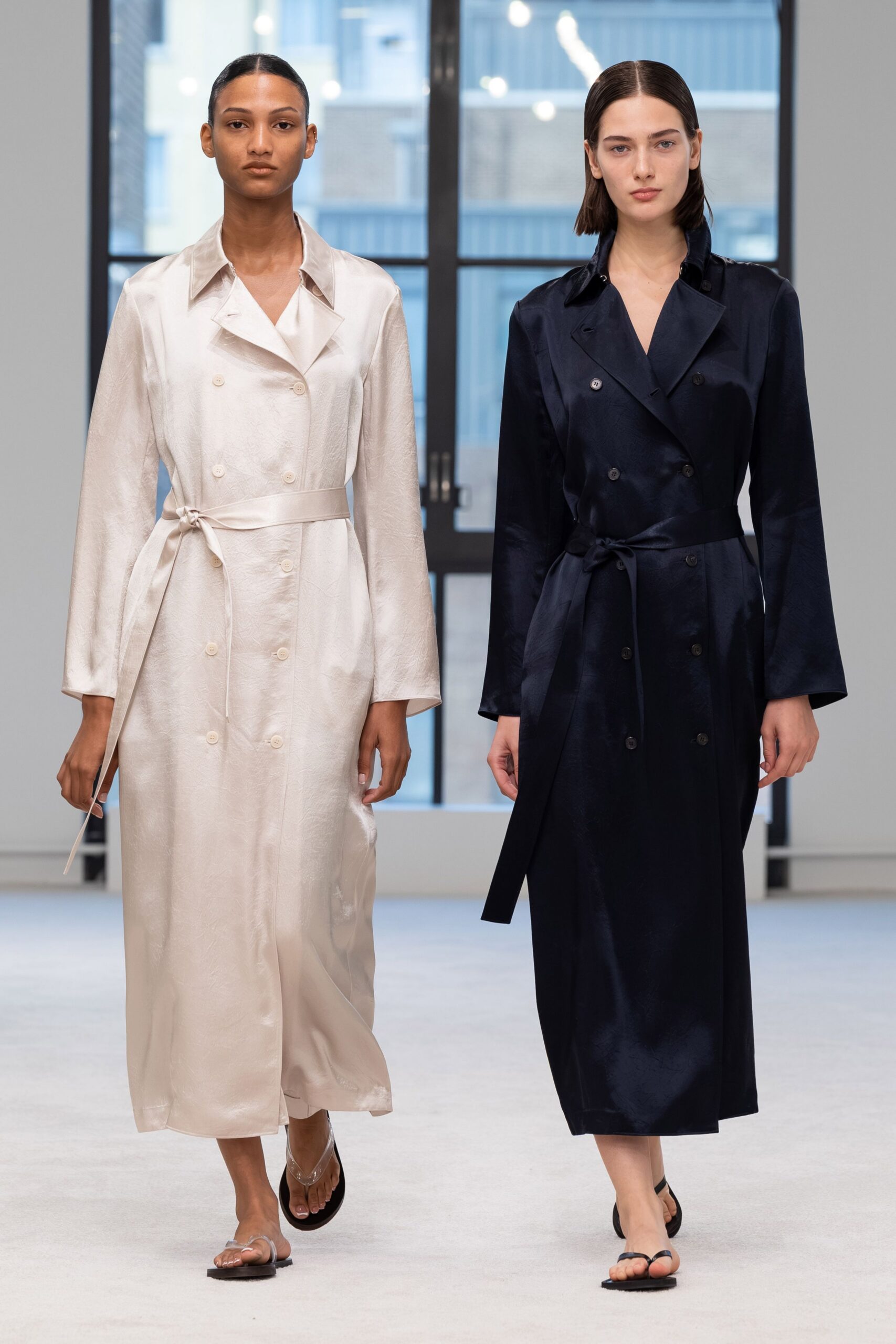 Ink satin twill coatdress, double-breasted with a self-tie hanging loose. Black thong flats. Crossing a quiet hotel atrium before breakfast service starts.