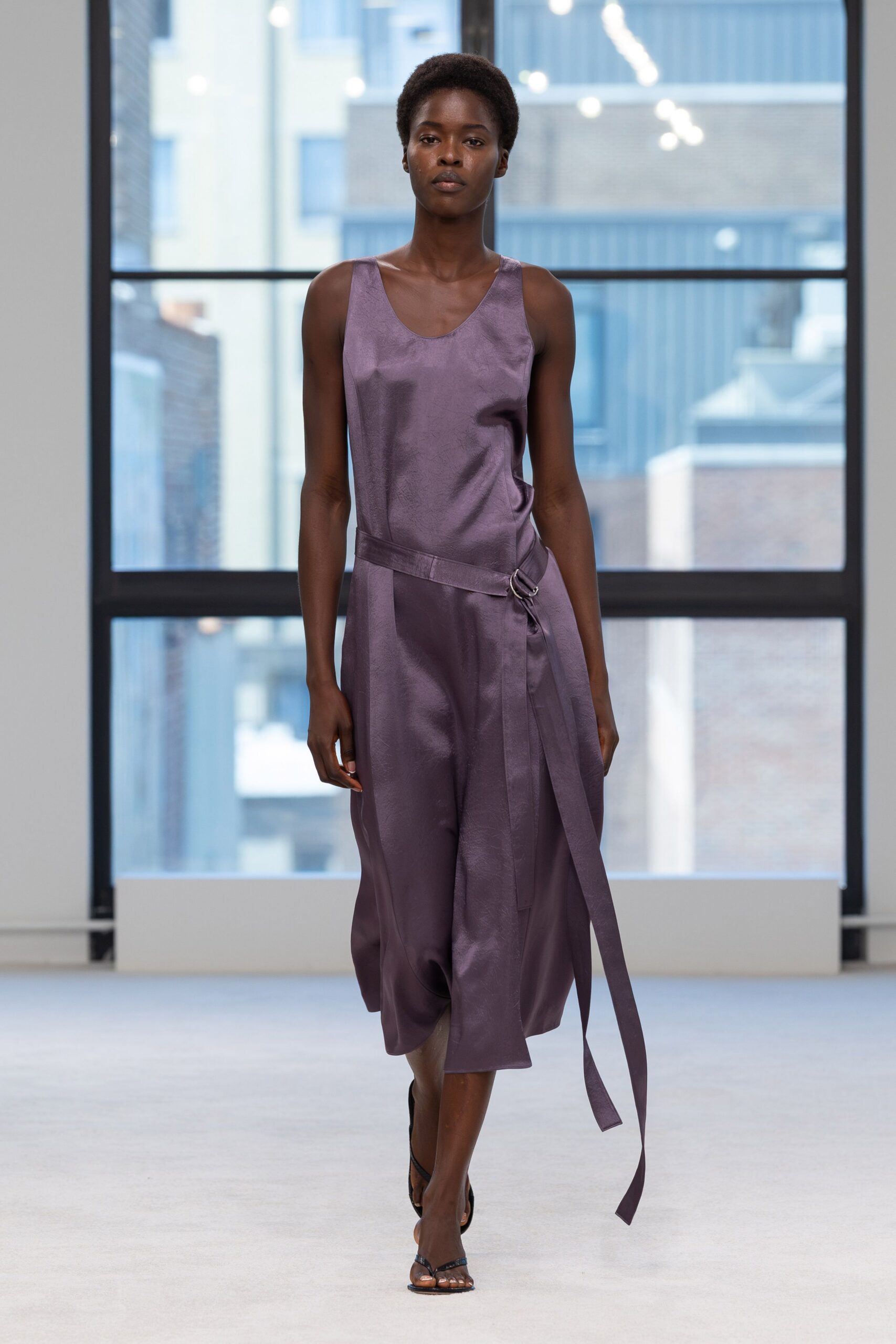 Mauve satin slip dress with a low scoop neck and wrap panel. Self-fabric belt looped through a silver D-ring, tail hanging to the calf. Black barely-there sandals. Crossing a glass atrium before the office cleaners have finished.