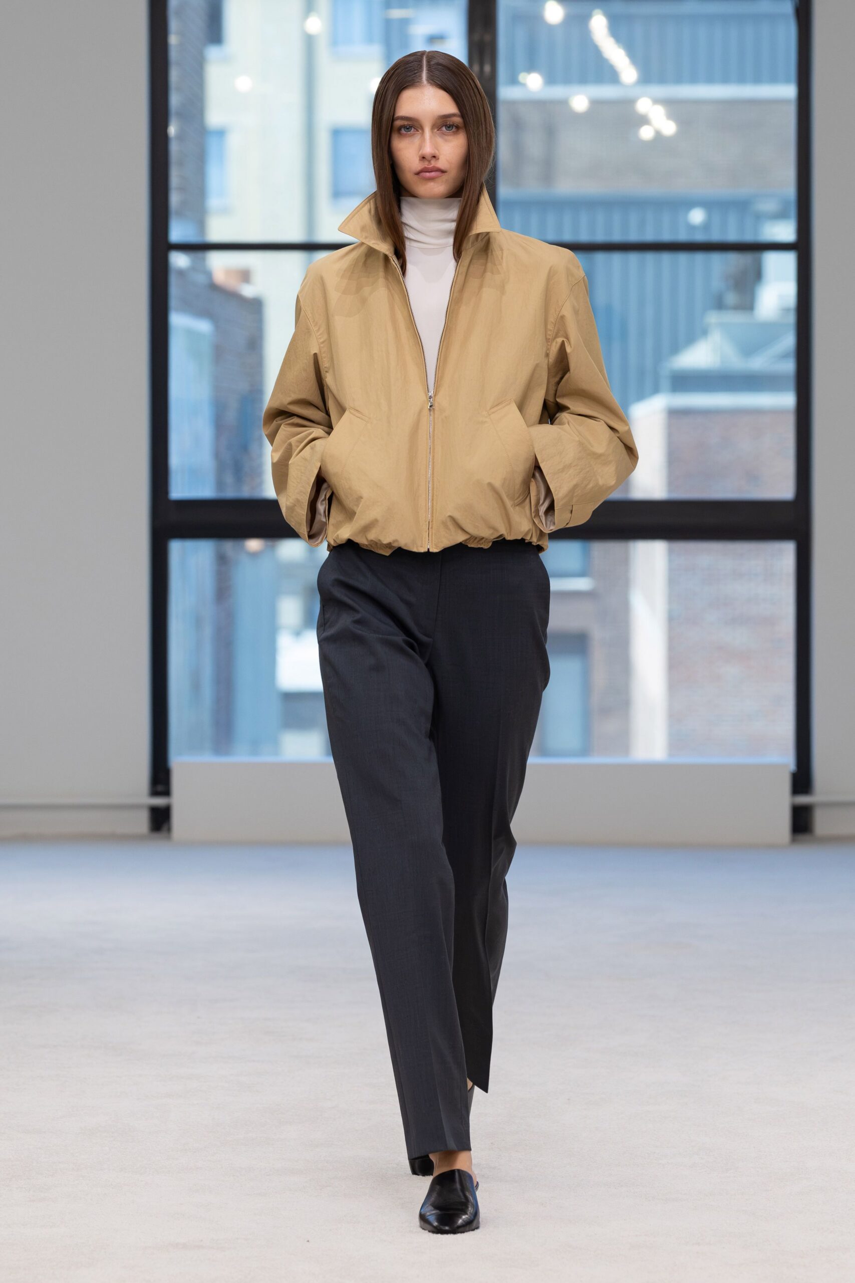 Sand cotton blouson with a standing collar and gathered hem. White jersey funnel-neck layer, smooth and close to the throat. Charcoal straight trousers with a crisp crease and slight break. Black leather mules with squared vamps. Early train to the office district before anyone has bought coffee.