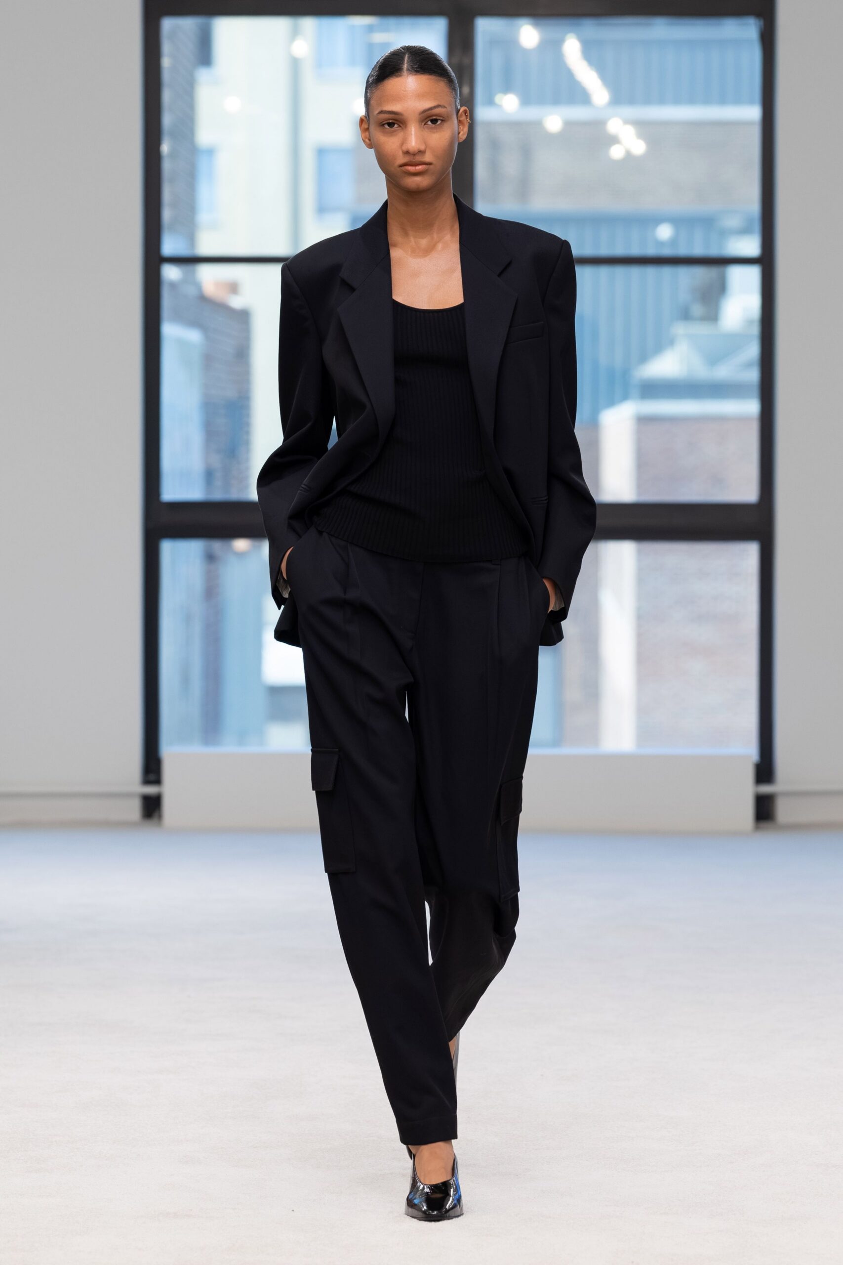 Black tailored blazer with broad peak lapels and a low single button. Black rib-knit tank, square neckline cut close across the chest. Black wool cargo trousers with flap pockets set low on the thighs. Black patent pumps with sharply pointed toes. First meeting in a glass tower, rain still drying on the pavement.