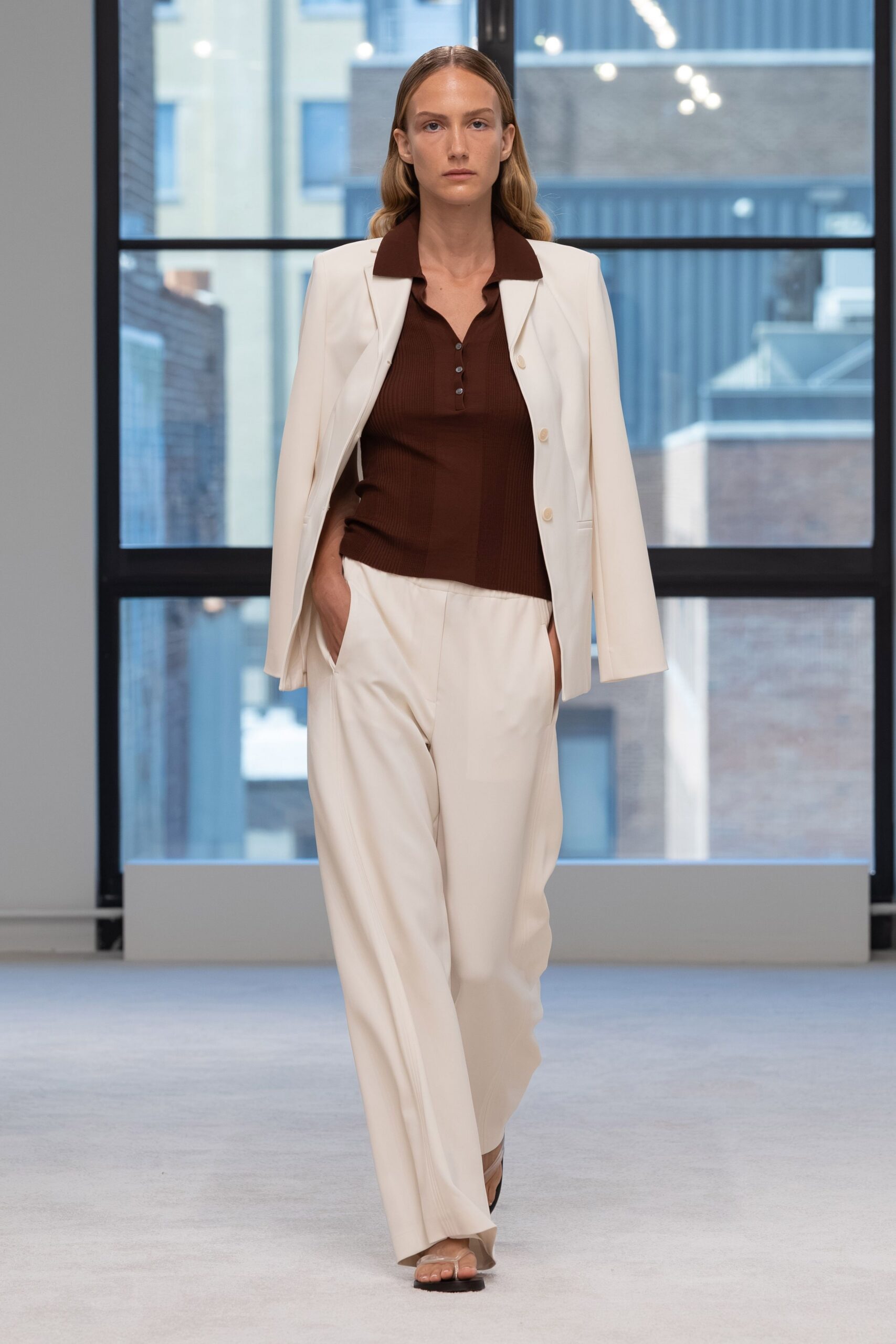 Cream blazer worn cape-like off the shoulders, matte buttons spaced low. Chocolate ribbed knit polo with a narrow four-button placket. Ivory fluid trousers falling straight from a clean flat front. Barely there thong sandals. First meeting in a glass office before the air conditioning kicks in.