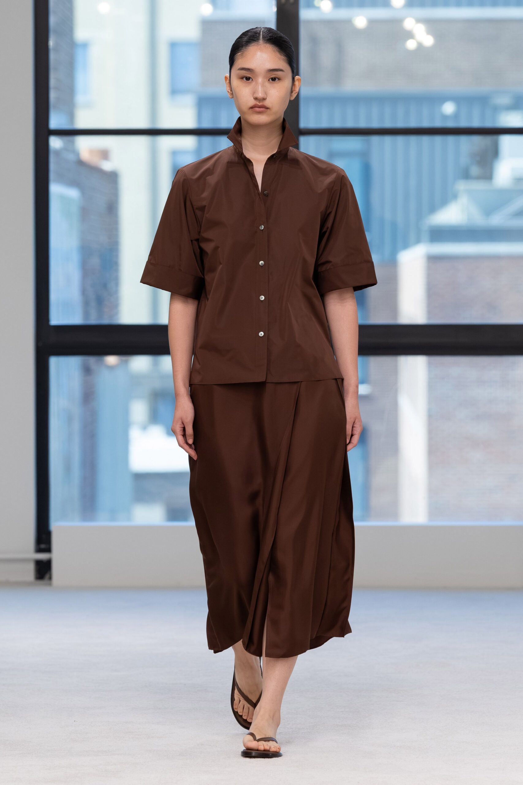 Chocolate poplin shirt with a high stand collar and turned-back cuffs. Matching satin wrap skirt, draped into a soft center fold. Flat brown thong sandals. Crossing a glass atrium before the office air-conditioning has caught up.