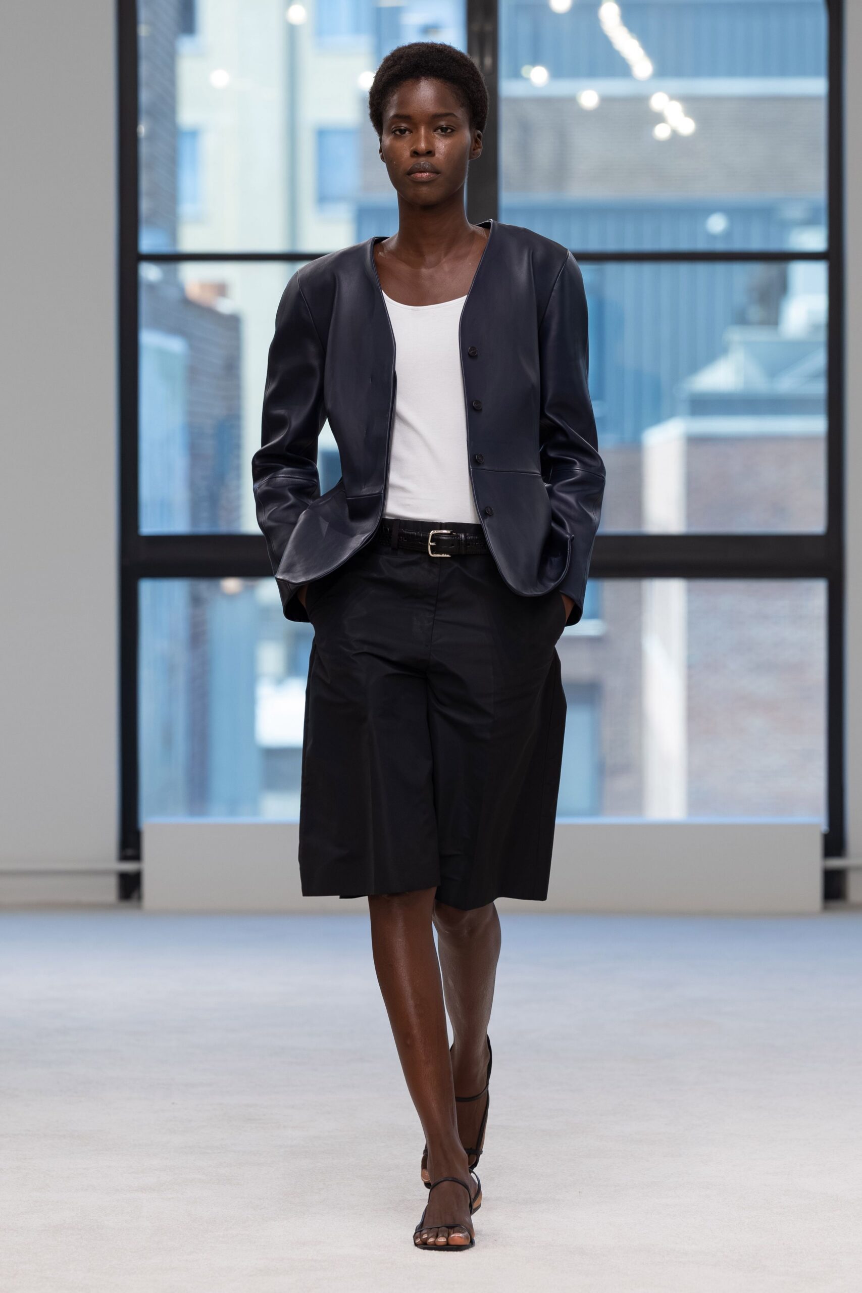 Ink leather jacket with a collarless edge and low rounded hem. White jersey tank cut in a wide scoop. Black tailored bermudas breaking just below the knee. Black leather belt with a square silver buckle. Bare black sandals with a slim toe strap. For crossing a glass office lobby before the first meetings start.