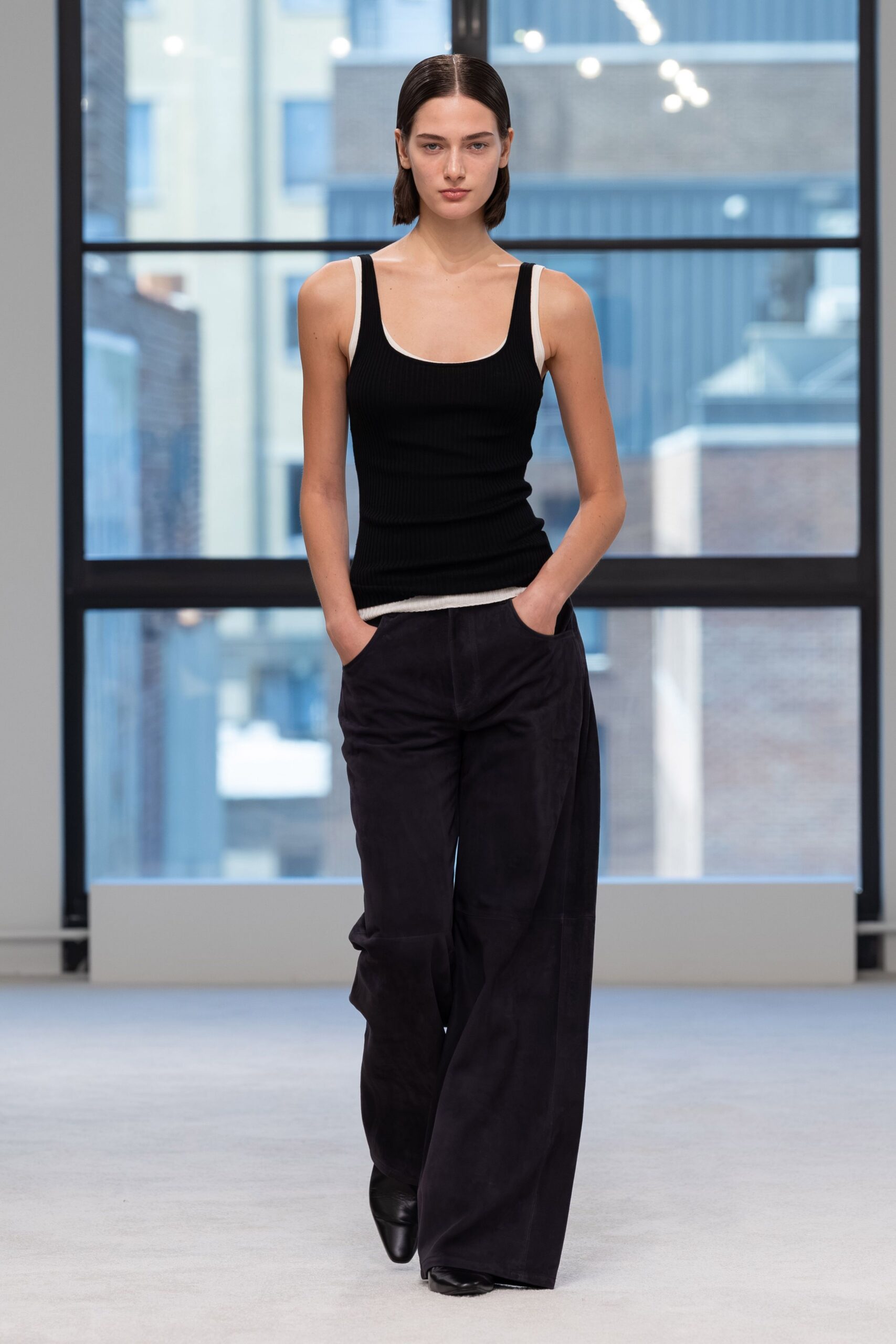 Black ribbed knit tank with white binding tracing the scoop neck. White jersey straps and hem peeking past the black layer. Ink-dark wide trousers in suede or brushed twill, hands sunk in pockets. Black pointed boots barely breaking the hem. Early office on a Saturday, city windows still blue with morning.