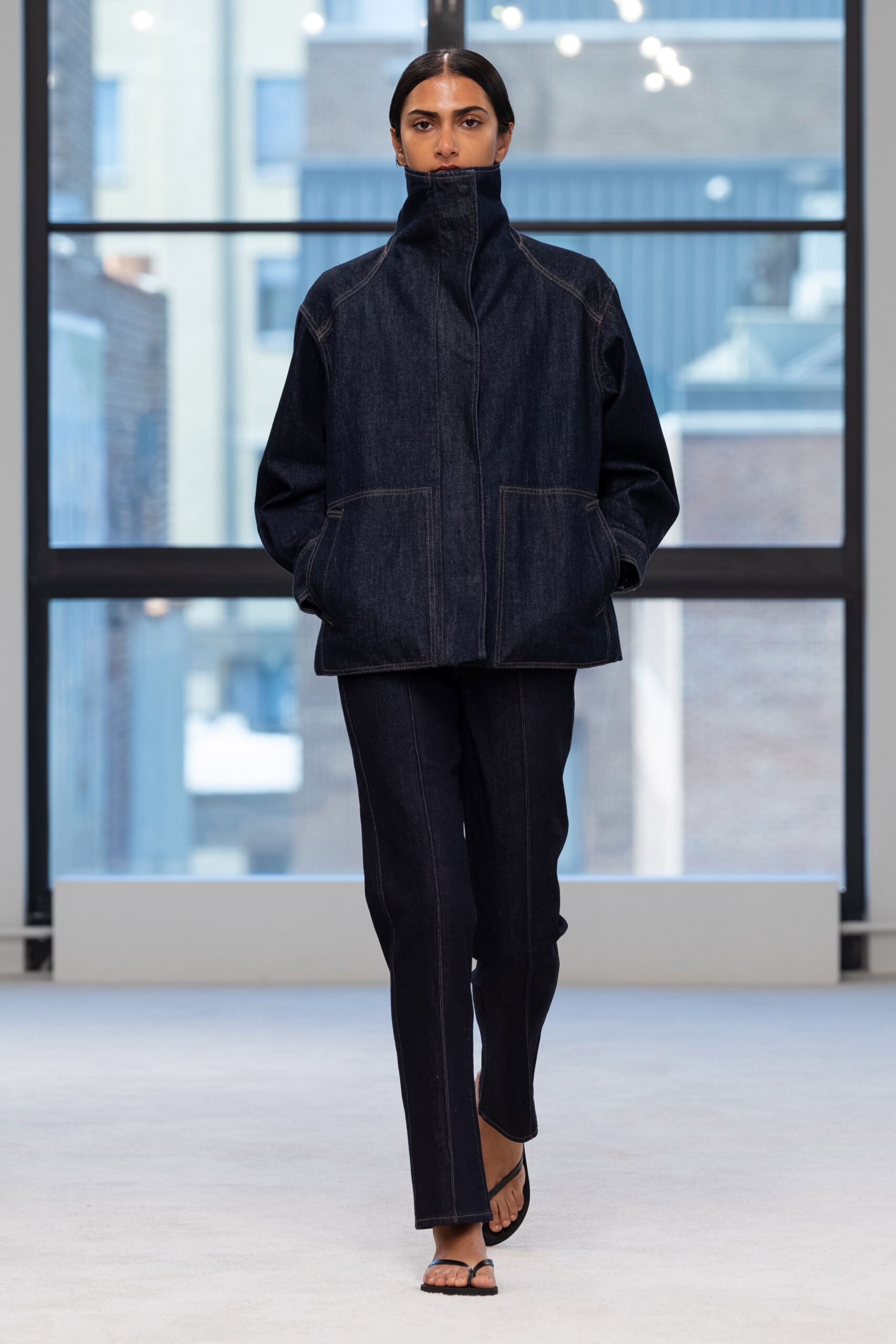 Dark indigo denim jacket with a zip hidden under the raised funnel neck. Matching straight-leg jeans, pressed by contrast topstitching down the front. Black thong sandals, barely there straps. Early train to the design office, coffee cooling in a paper cup.