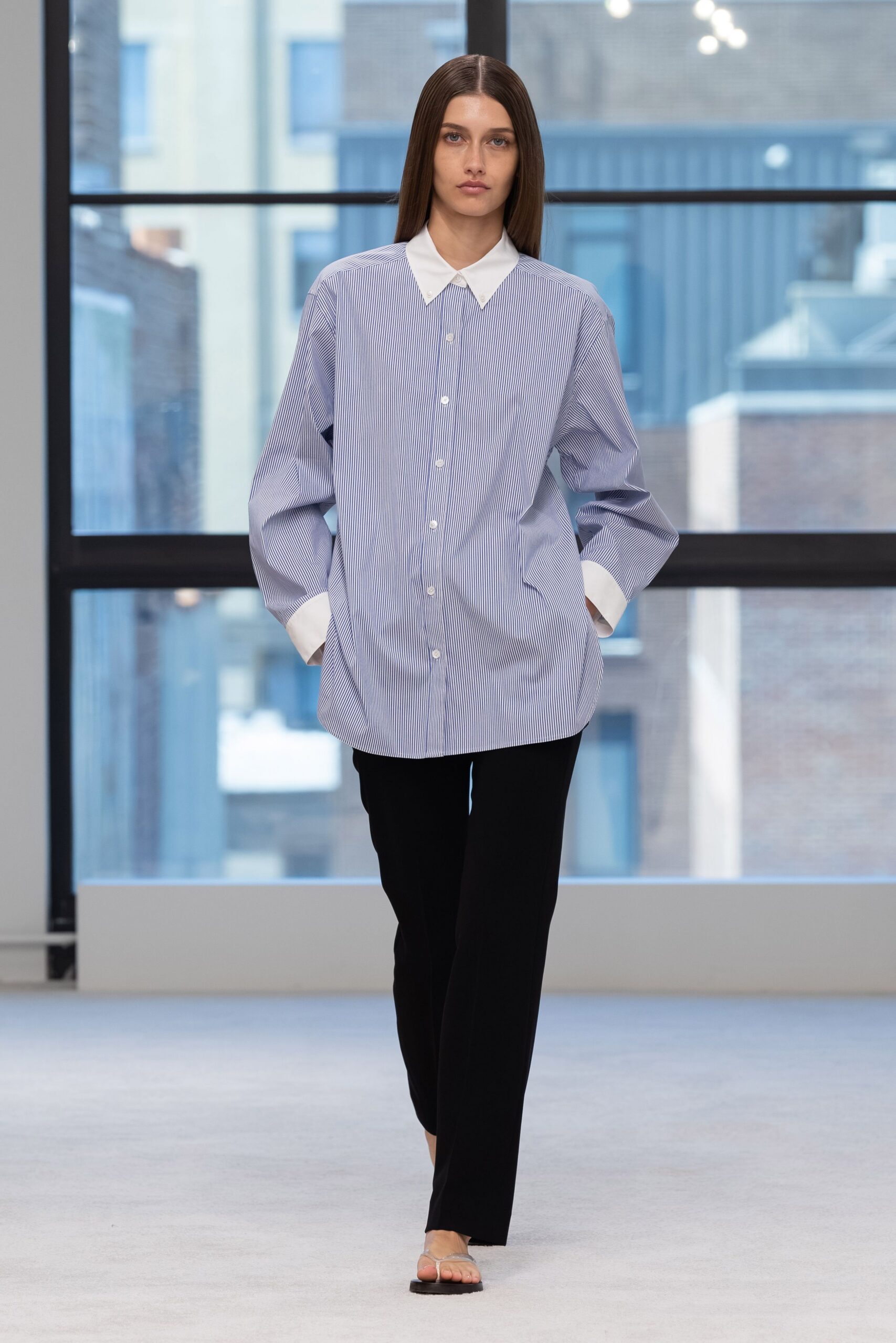 Oversized blue banker-stripe shirt with a stark white collar and deep cuffs. Black straight trousers falling cleanly from the hip. Barely-there thong sandals with clear straps over the toes. First meeting after moving offices, city roofs still blue through the glass.