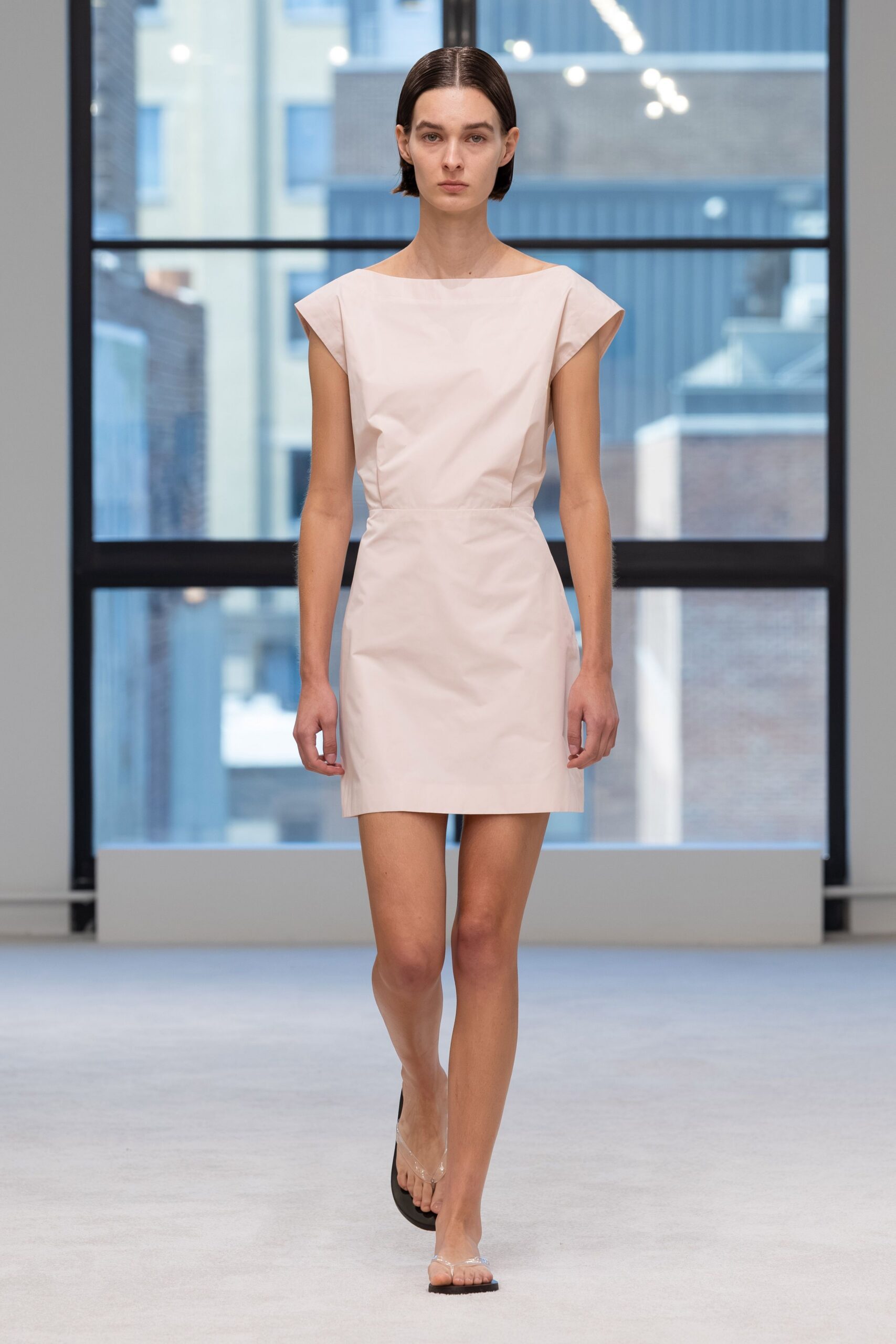 Pale shell-pink mini dress in crisp cotton, cut with cap sleeves and a boatneck. Clear thong sandals on flat black soles. Early gallery shift, windows still blue from the morning rain.