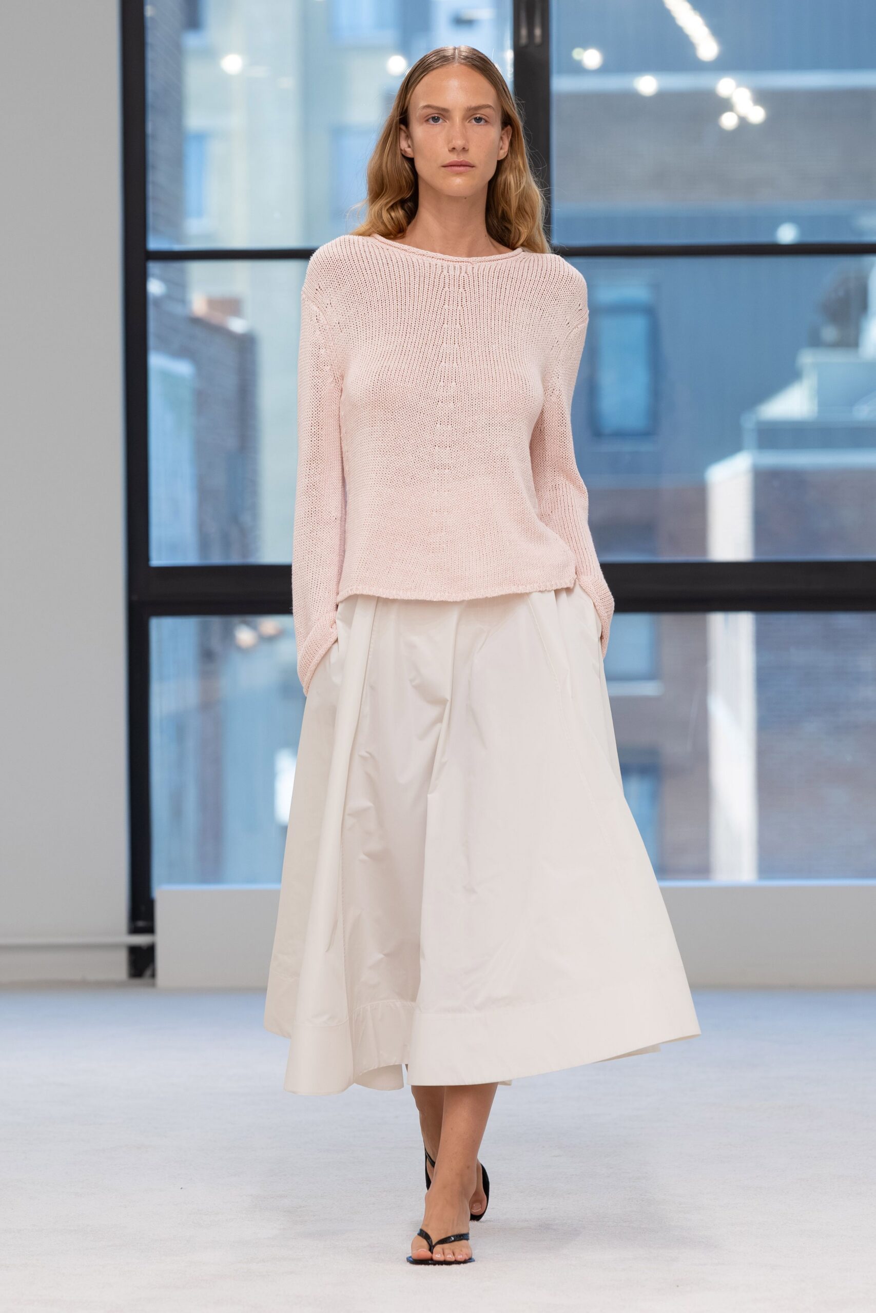 Blush open-knit sweater with a rolled boatneck and dropped shoulders. Ivory cotton poplin midi skirt, cut full with soft collapsed volume. Black thong sandals with thin patent straps. Windowlight in a borrowed apartment, the city still blue before dinner.