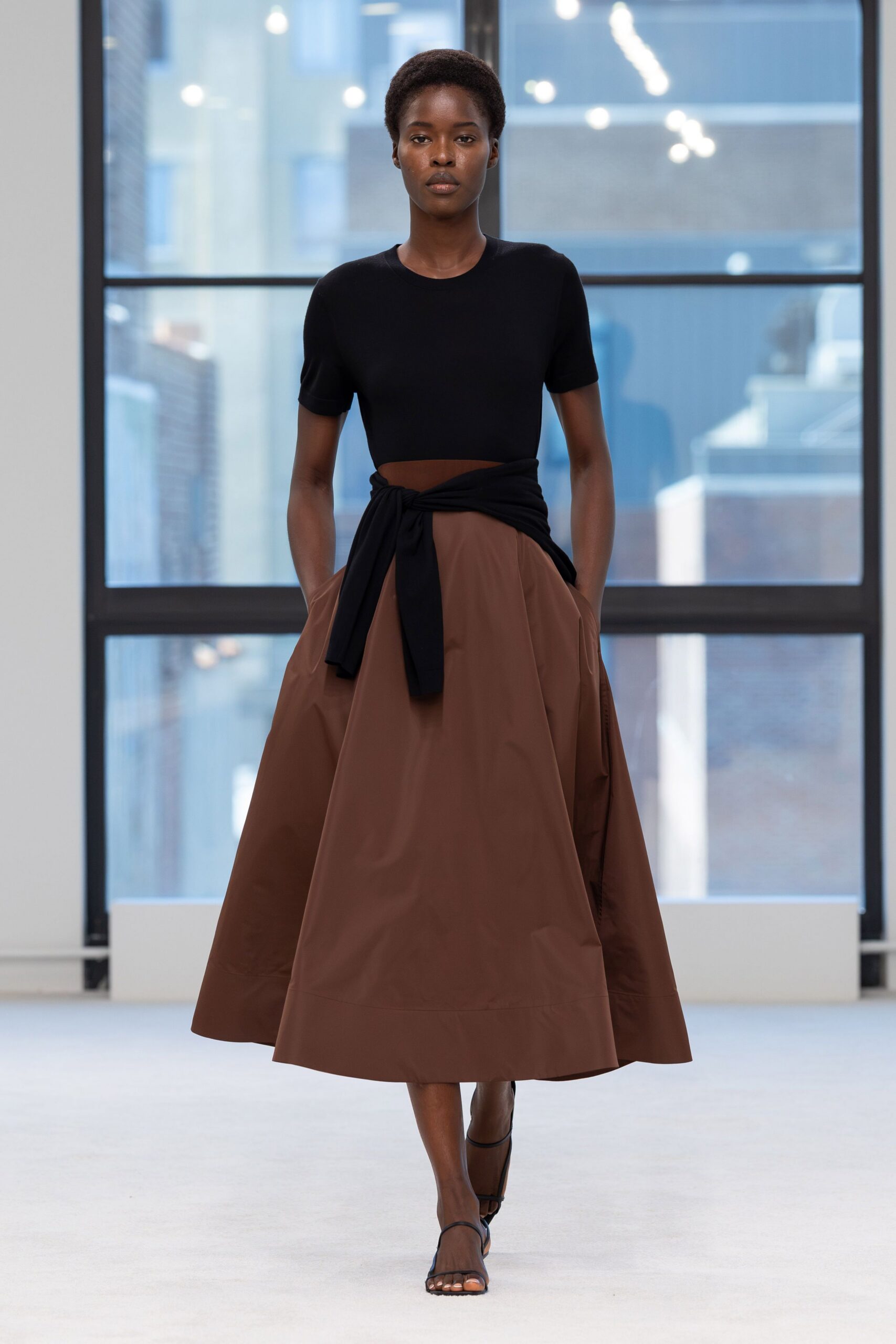 Black fine-knit cropped top with a clean crew neck and short sleeves. Chestnut paper-taffeta skirt ballooning to mid-calf, hands sunk in side pockets. Black jersey panel knotted low, leaving a crescent of bare midriff. Black barely-there ankle-strap sandals. Office tower atrium, the hour before anyone else arrives.