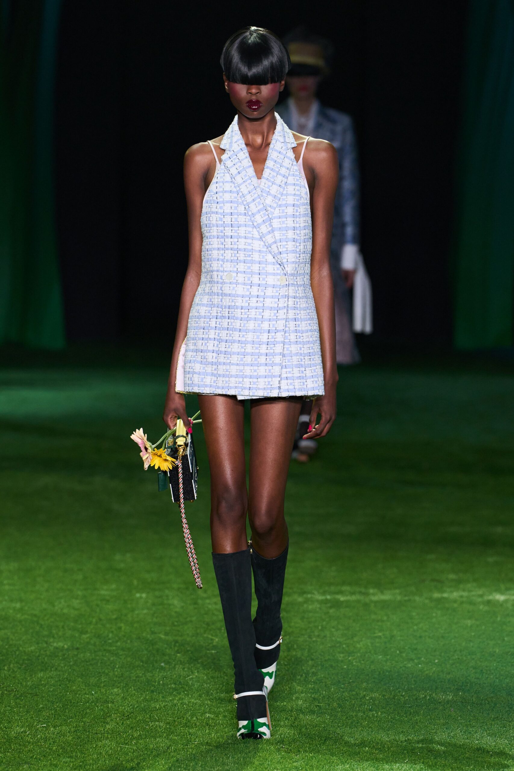 Powder-blue tweed halter playsuit cut like a double-breasted blazer. Box bag with a braided shoulder cord and fresh daisies tucked in. Black ribbed leg warmers to mid-calf. White heels with green splash print and a thin ankle strap. Crossing a lawn after brunch, still holding the flowers from the table.