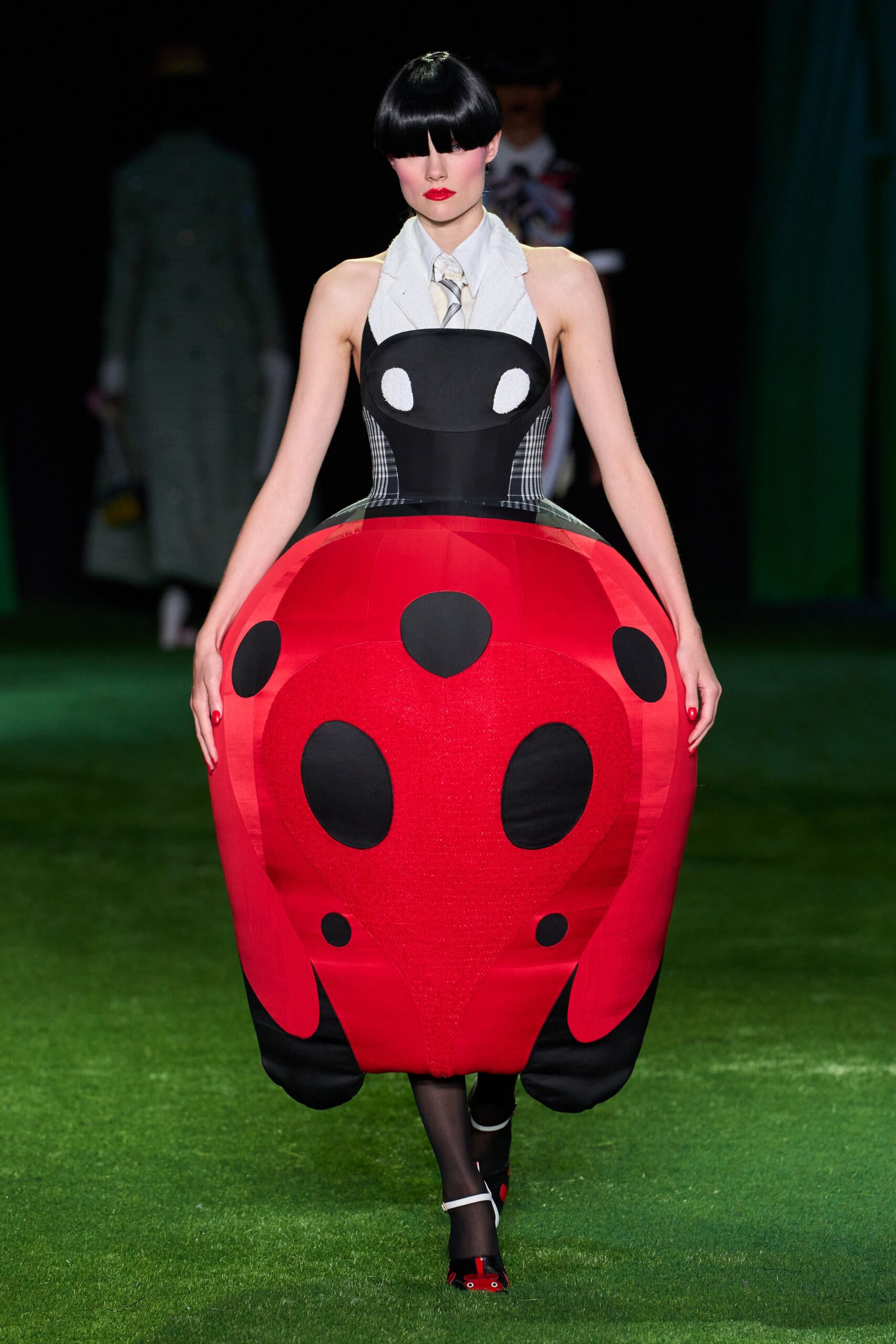 Pale grey shirt collar and striped tie peeking through the halter opening. Ladybird cocoon dress, padded scarlet shell with black spots and plaid side panels. Sheer black tights. White-strap heels with tiny cartoon eyes at the toe. Crossing a miniature-golf green after the children's party emptied out.