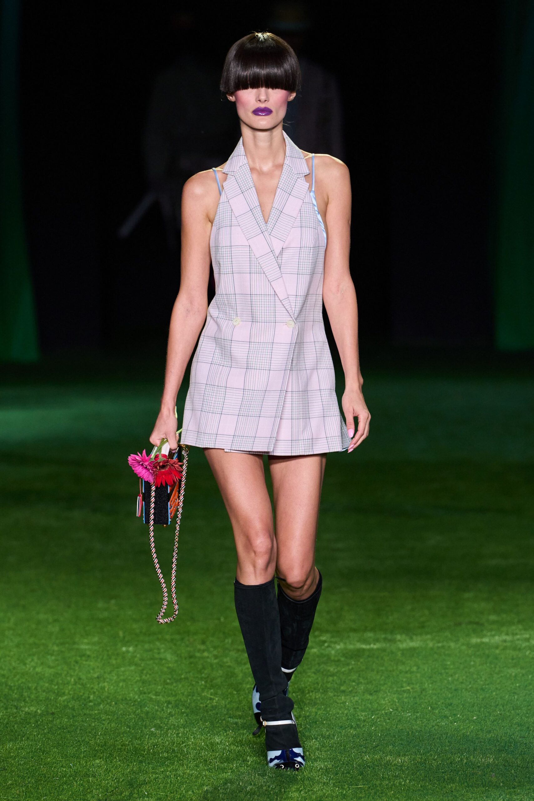 Powder-pink plaid mini cut like a double-breasted blazer, blue straps showing. Small woven box bag with braided cord strap and two pinned gerberas. Black slouch boots over mismatched blue-and-white pointed shoes. Walking across wet grass after a garden wedding everyone stayed too long at.