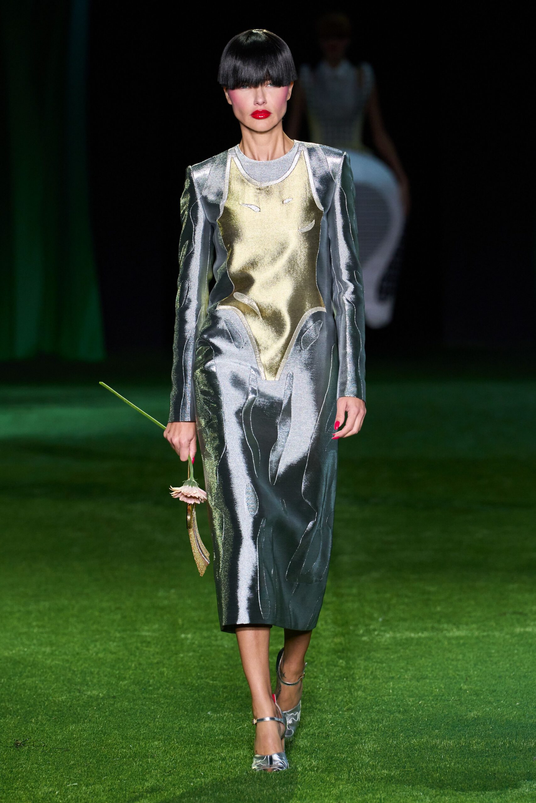 Liquid silver sheath with a pale gold torso panel and raised contour seams. Heather-grey crewneck knit peeking cleanly at the collar. Gold mesh sling bag carrying a drooping pink gerbera. Silver ankle-strap pumps with translucent pointed vamps. Crossing a wet lawn after the wedding, one flower already giving up.