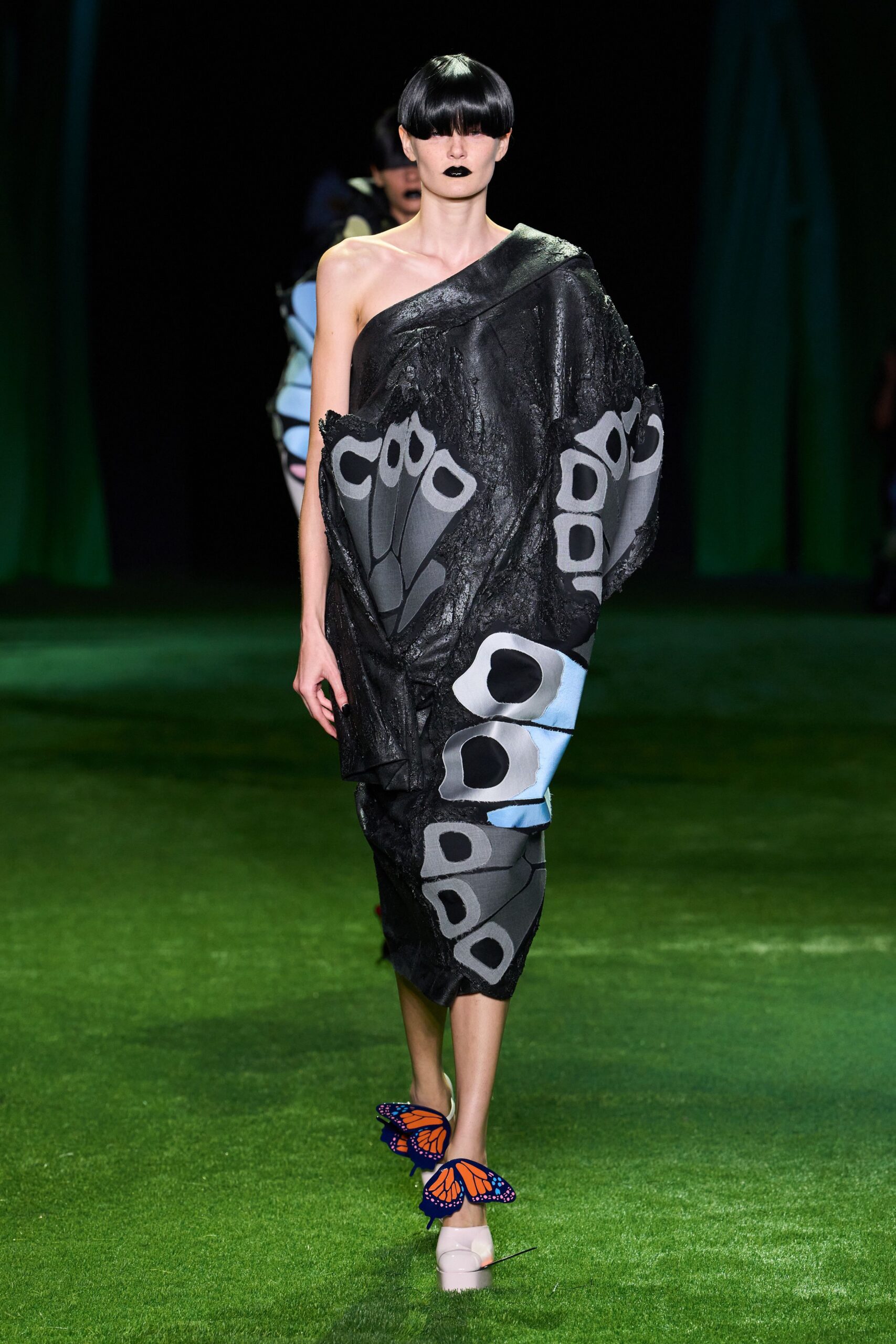 One-shoulder black cocoon dress in crinkled coated fabric, moth-wing panels wrapping the body. Pale platform heels with monarch butterflies clipped over the toes. Crossing a museum lawn after dark while the insect exhibit is still humming.