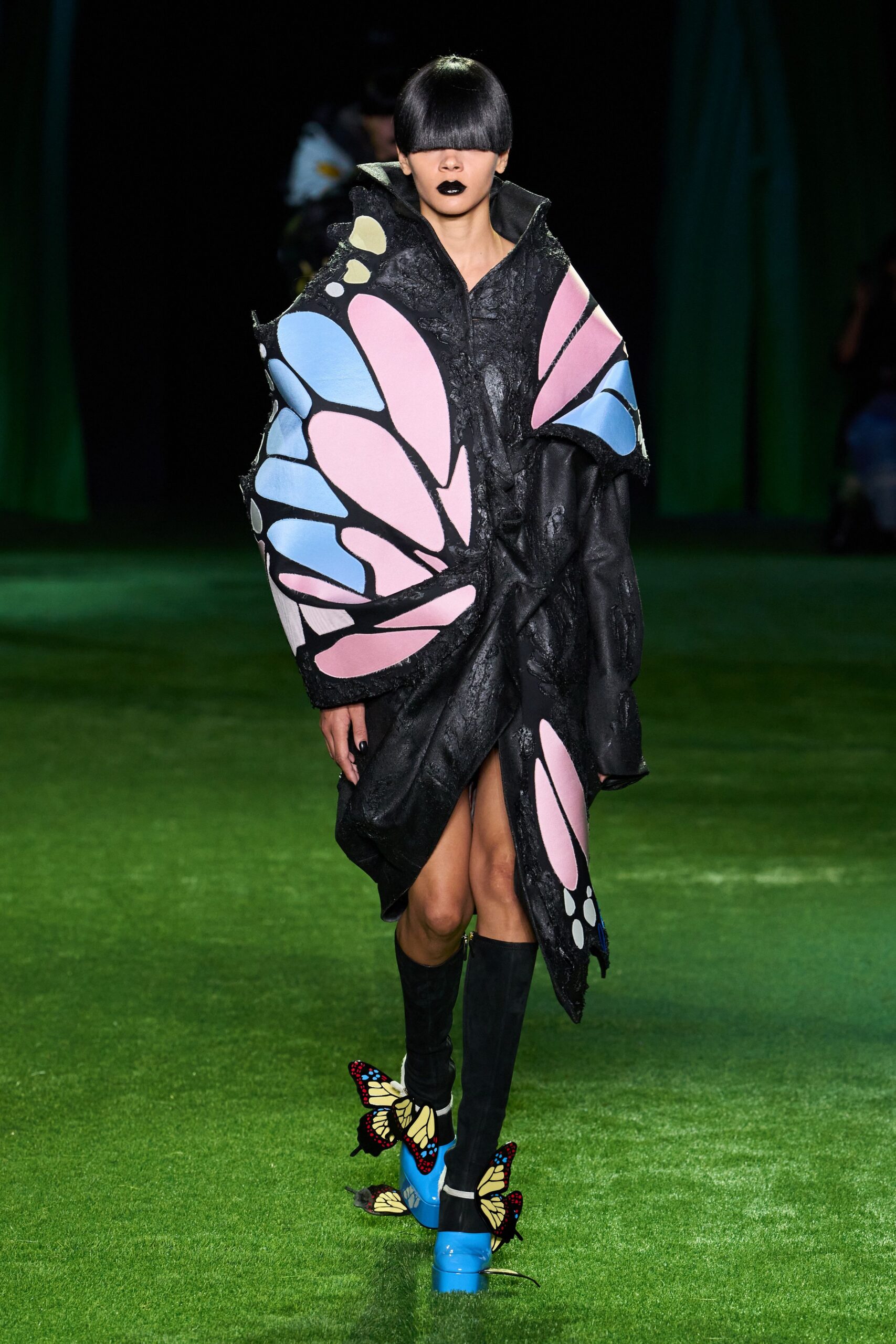 Black sculpted coat dress with raised collar and pink-blue butterfly panels. Plain black knee socks. Sky-blue patent platforms with butterfly wings fluttering off the ankles. Crossing wet grass after dusk, still wearing the costume from the garden maze.