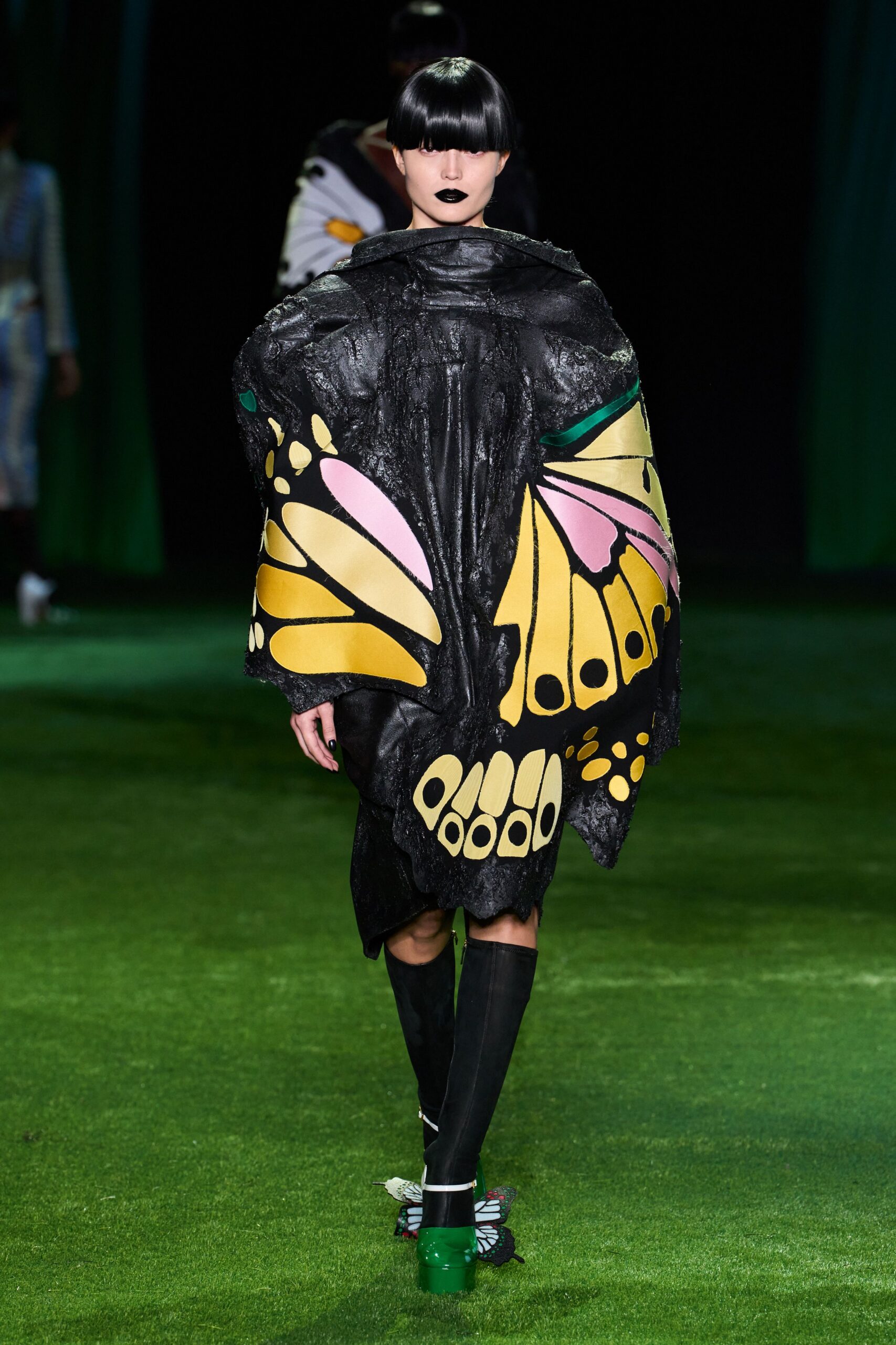 Crinkled black shell cape painted with oversized butterfly wings in lemon and pink. Matching black skirt with a ragged hem peeking below. Opaque black knee socks. Green patent platforms with butterfly-shaped soles trailing behind. Crossing a museum lawn after dark, still wearing the installation.