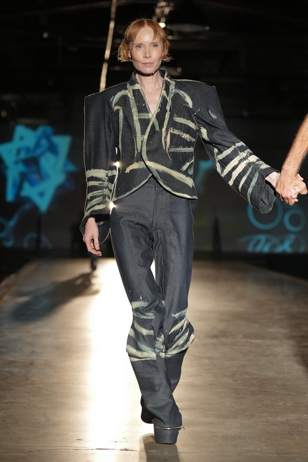Charcoal wrap jacket with bleached brushstrokes and cutaway slits under the shoulders. Matching denim trousers, pale streaks circling the lower legs. Black platforms with a rounded block sole. Crossing an empty gym floor after someone killed the lights.