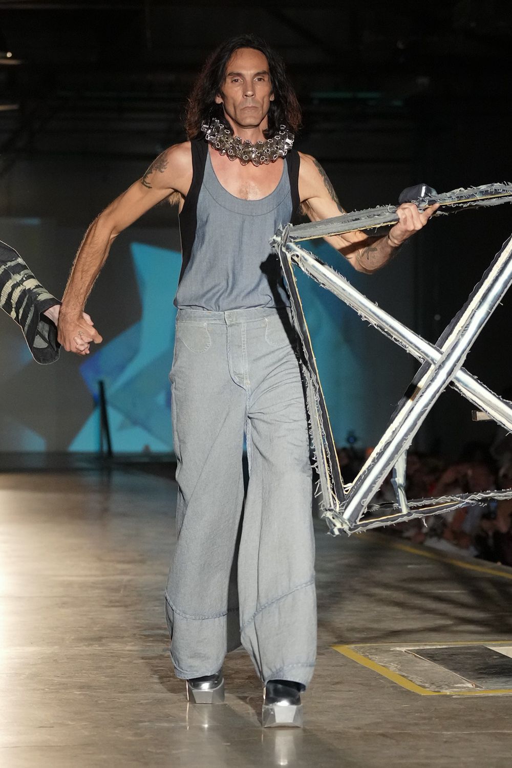 Clear plastic bottle tops clustered into a rattling collar. Washed slate tank with a scooped neckline and black side panels. Pale denim balloon trousers, curved seams scooping over the shins. Black shoes on mirrored block platforms. Crossing an empty loading bay carrying the stripped frame of a bicycle.