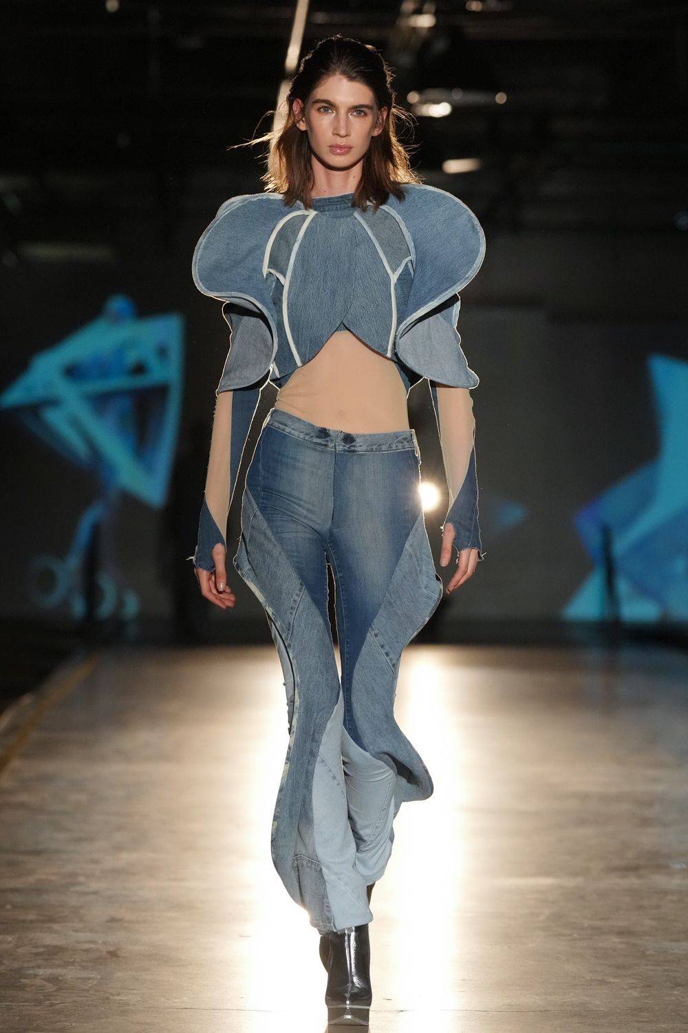 Blue chambray cropped top with swollen petal shoulders and white piped seams. Nude stretch bodysuit cut high across the bare midriff. Spliced denim trousers with long split panels whipping around the legs. Black ankle boots, sharply squared at the toe. Crossing an empty convention hall while the projector tests start up.