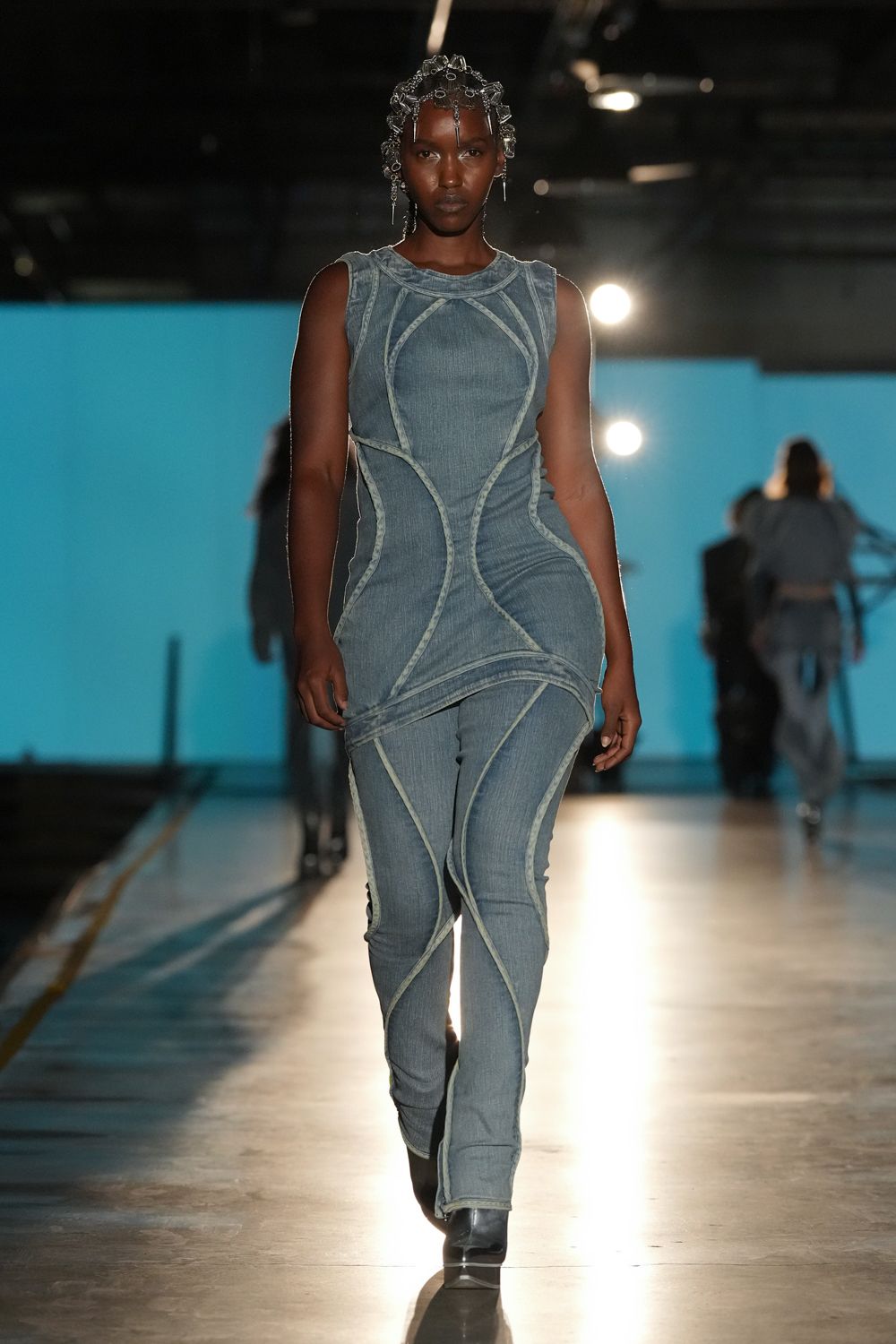 Crystal-beaded chain headpiece, one drop hanging between the brows. Faded blue ribbed denim tunic, pale curved seams mapping the torso. Matching denim leggings with looping flatlocked seams and split hems. Square-toe black boots. Crossing the station concourse after rain, lights glaring off the floor.