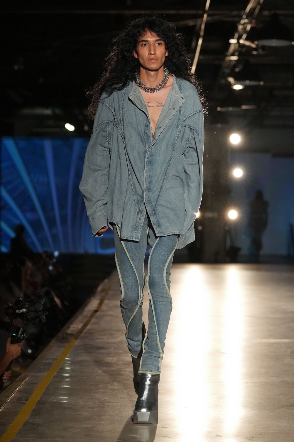 Heavy silver chain clustered tight at the collarbone. Washed blue chambray shirt, dropped seams and snap placket hanging open. Spray-tight faded jeans traced with pale curved piping down the legs. Black leather ankle boots with blunt square wedge toes. Walking the depot floor before sunrise, generators already humming.