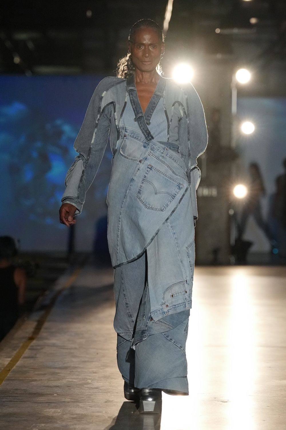 Washed pale denim wrap top with spliced sleeves and loose belt ends. Patchworked denim maxi built from jeans panels, back pocket swung to the front. Black platform boots with a blunt square toe. Crossing the fairgrounds before noon, generators already humming.